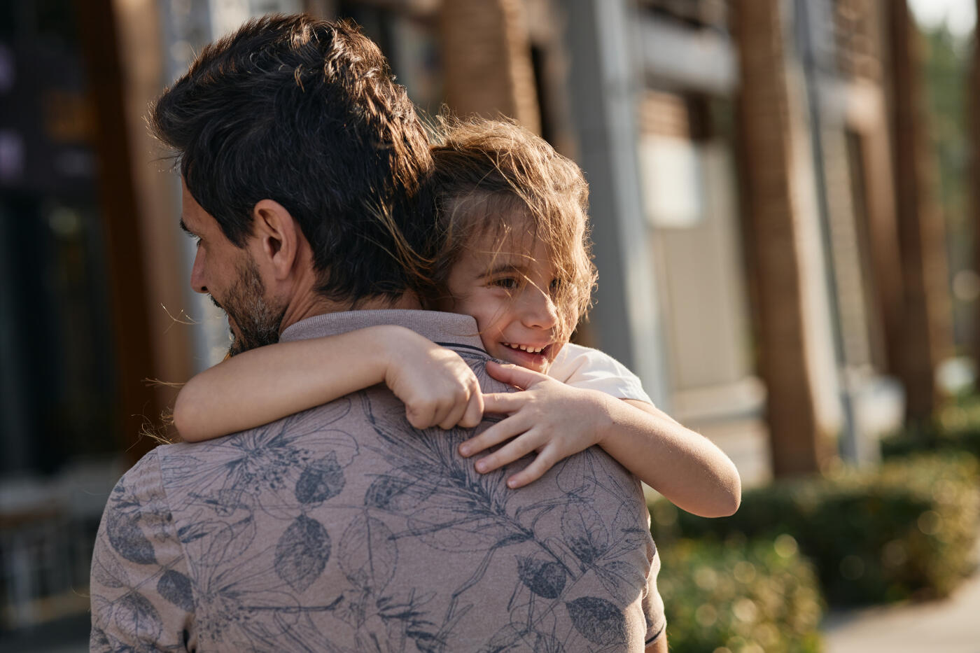 Attractive father carrying his beautiful daughter while walking through tropical streets with palm trees. They are happy and smiling.
