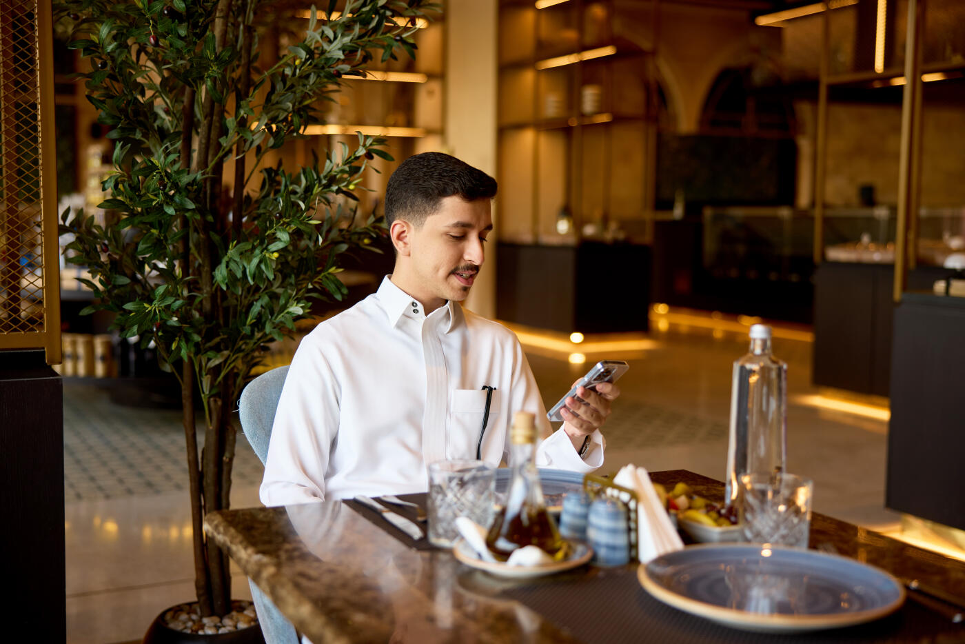 Waist up view of traditionally dressed individual in early 20s sitting at table and smiling while using smart phone.