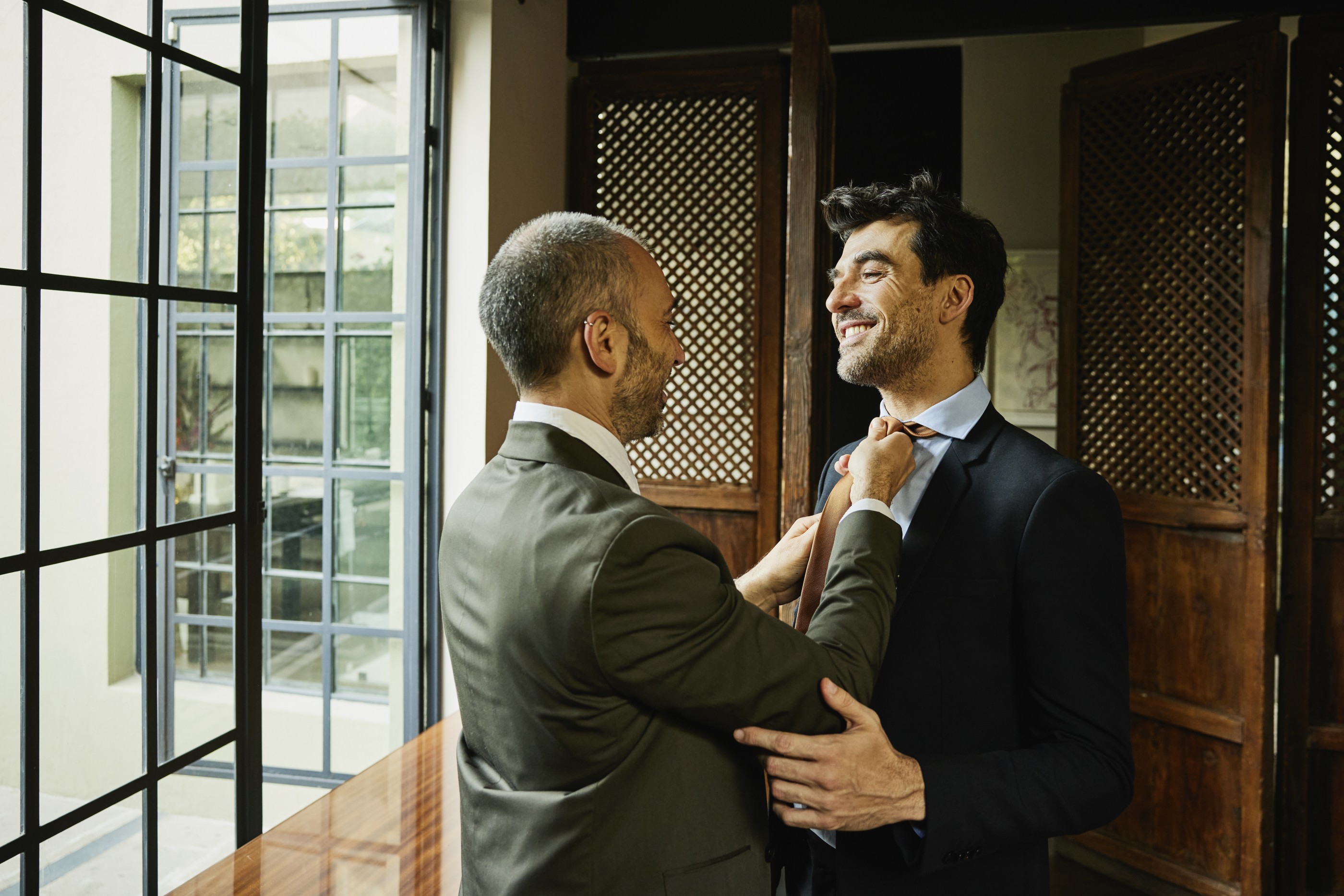 Medium shot of smiling man helping husband tie necktie in bedroom of boutique hotel before going out for evening