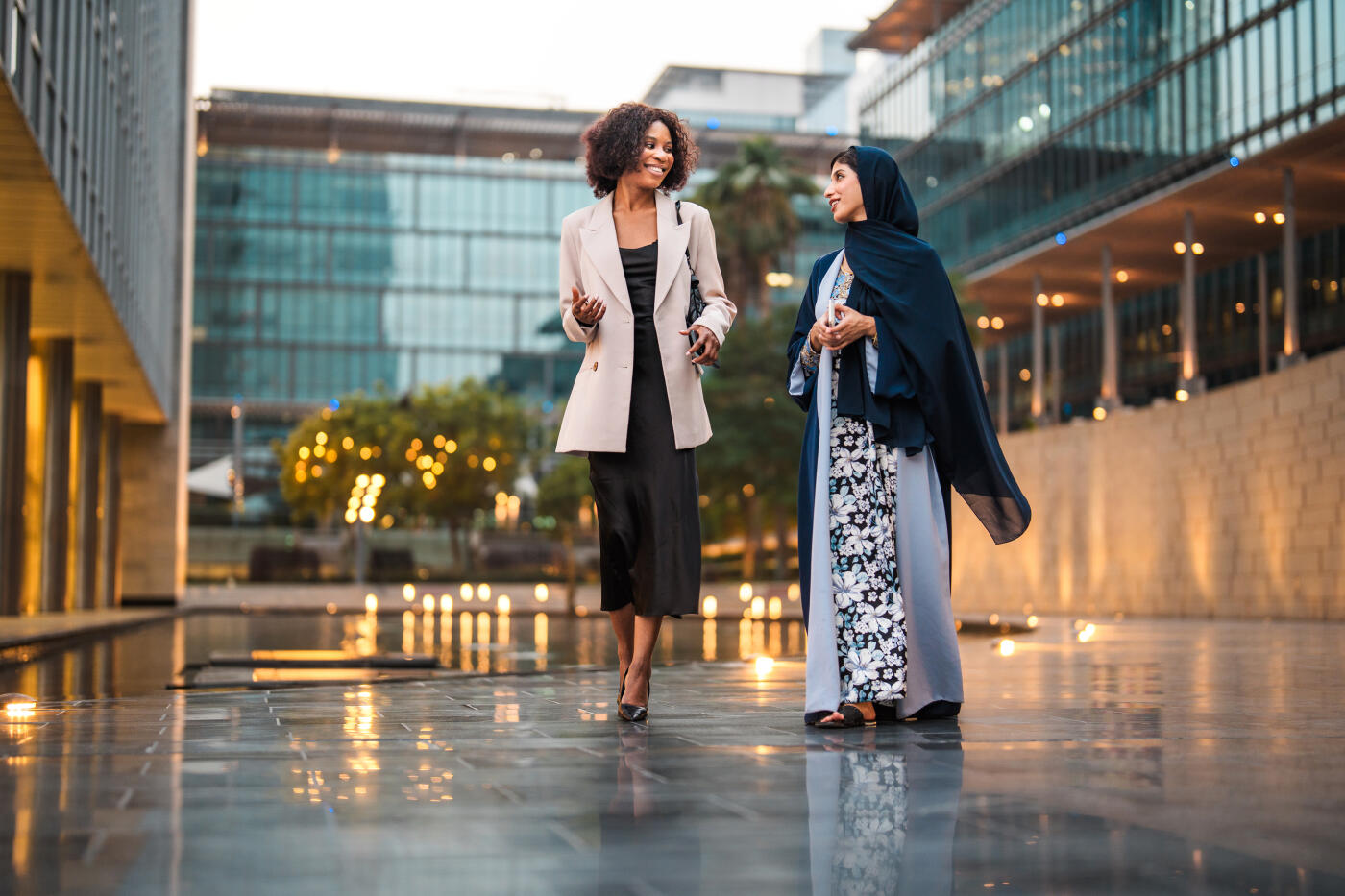 Two women engaging in conversation while walking through a modern financial district plaza. Glass and steel buildings frame the scene, with warm golden hour lighting reflecting off wet pavement. Professional attire and animated gestures convey active dialogue.