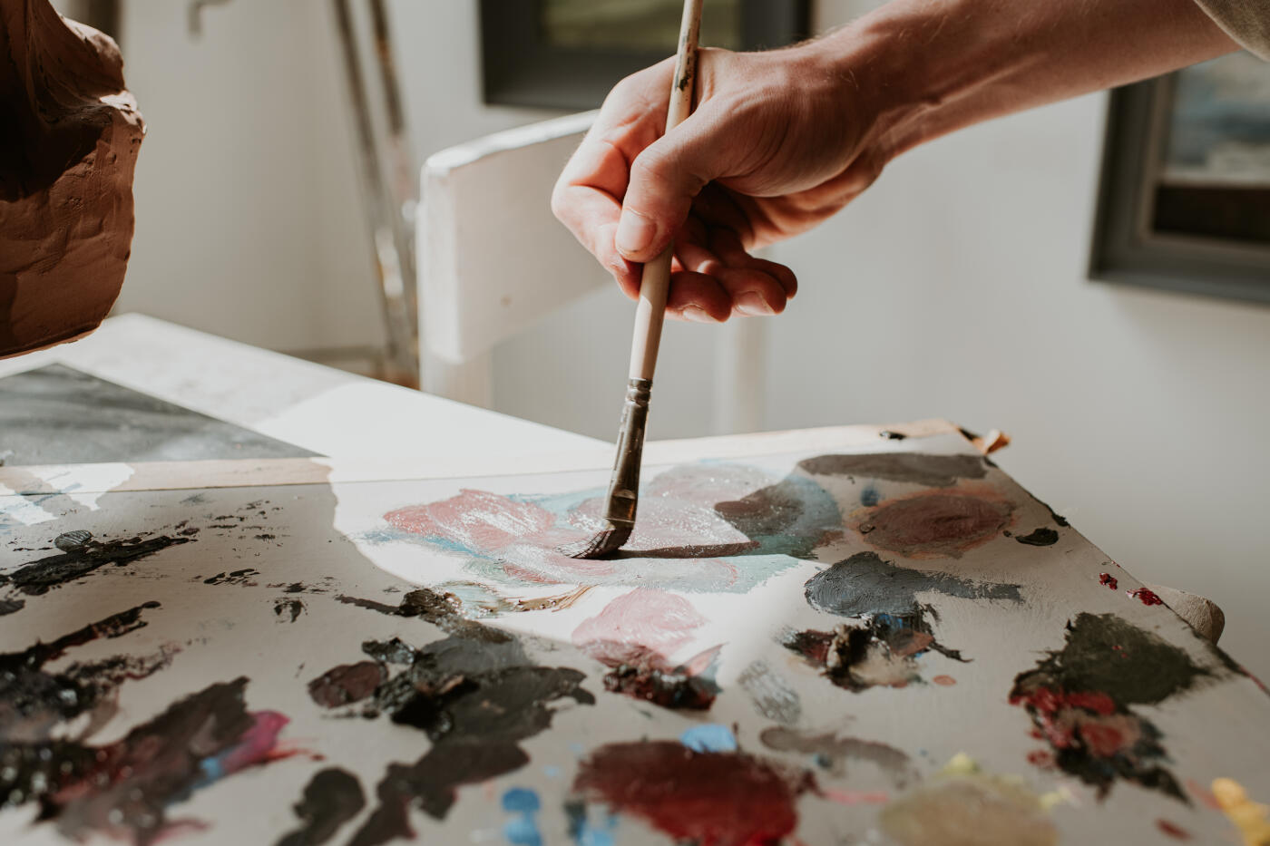 Sun illuminates a surface with wet oil paint. A hand holds a paint brush and mixes colours.