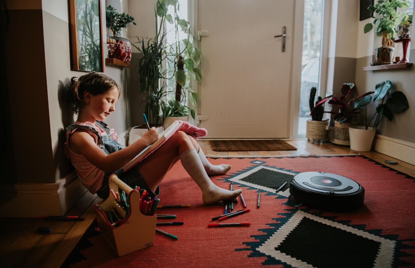 A little girl sits on the floor, concentrating on her artwork, doodling in a notebook with felt tip pens. The pens lie all over the floor around her. A robot vacuum cleaner quietly and efficiently works around her, cleaning the carpet.