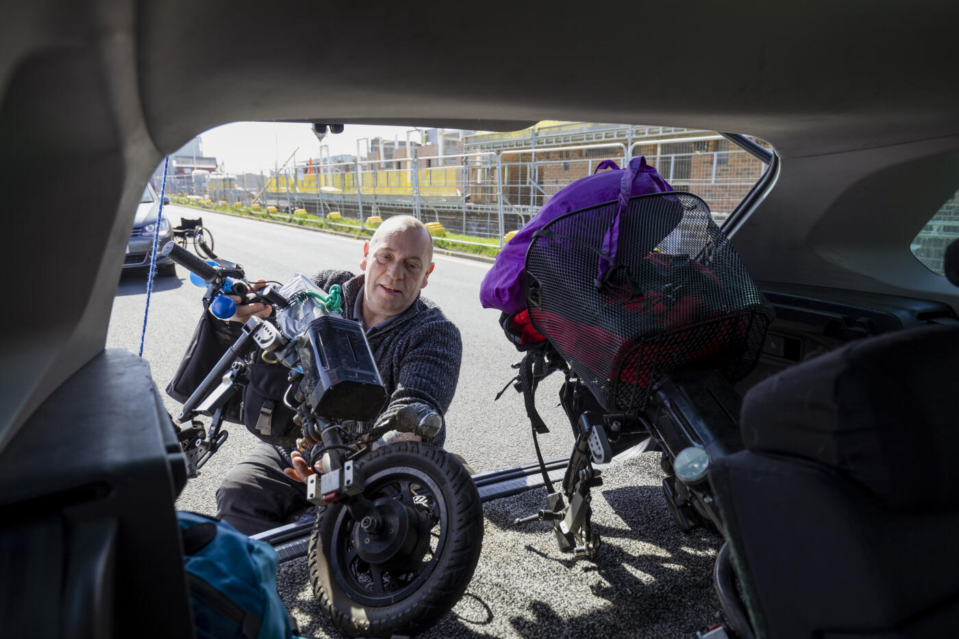 A front view of a mature man unloading the motorized part of his wheelchair out of an adapted car trunk. His car trunk is adapted to accommodate the attachments which motorize his wheelchair. He is arriving in Blyth to meet his friends for a day out fishing.