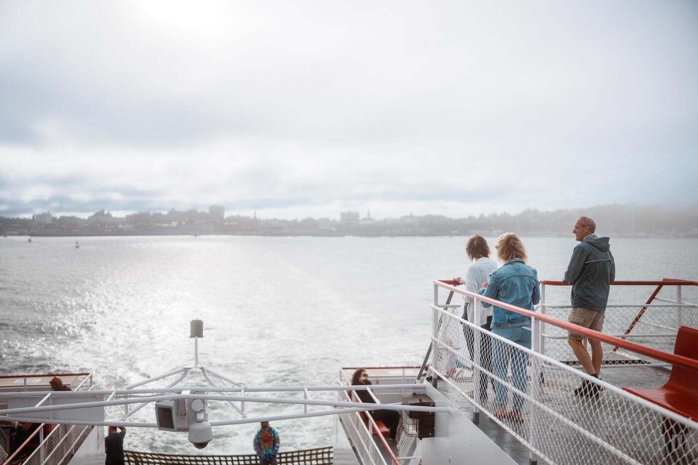Three senior friends stand on the deck of a ferry looking back at the Portland, Maine skyline in the distance while traveling to Peaks Island.