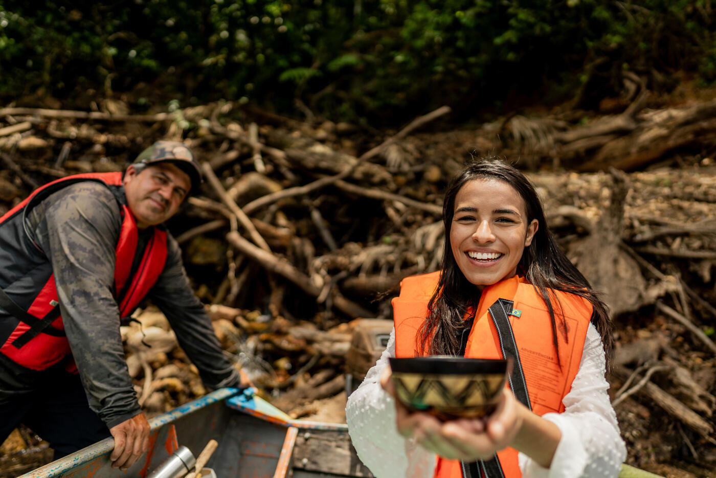 A young woman wearing a life jacket smiles brightly while holding a colorful handmade bowl, with a man standing on a boat and a background full of tree roots and forest.