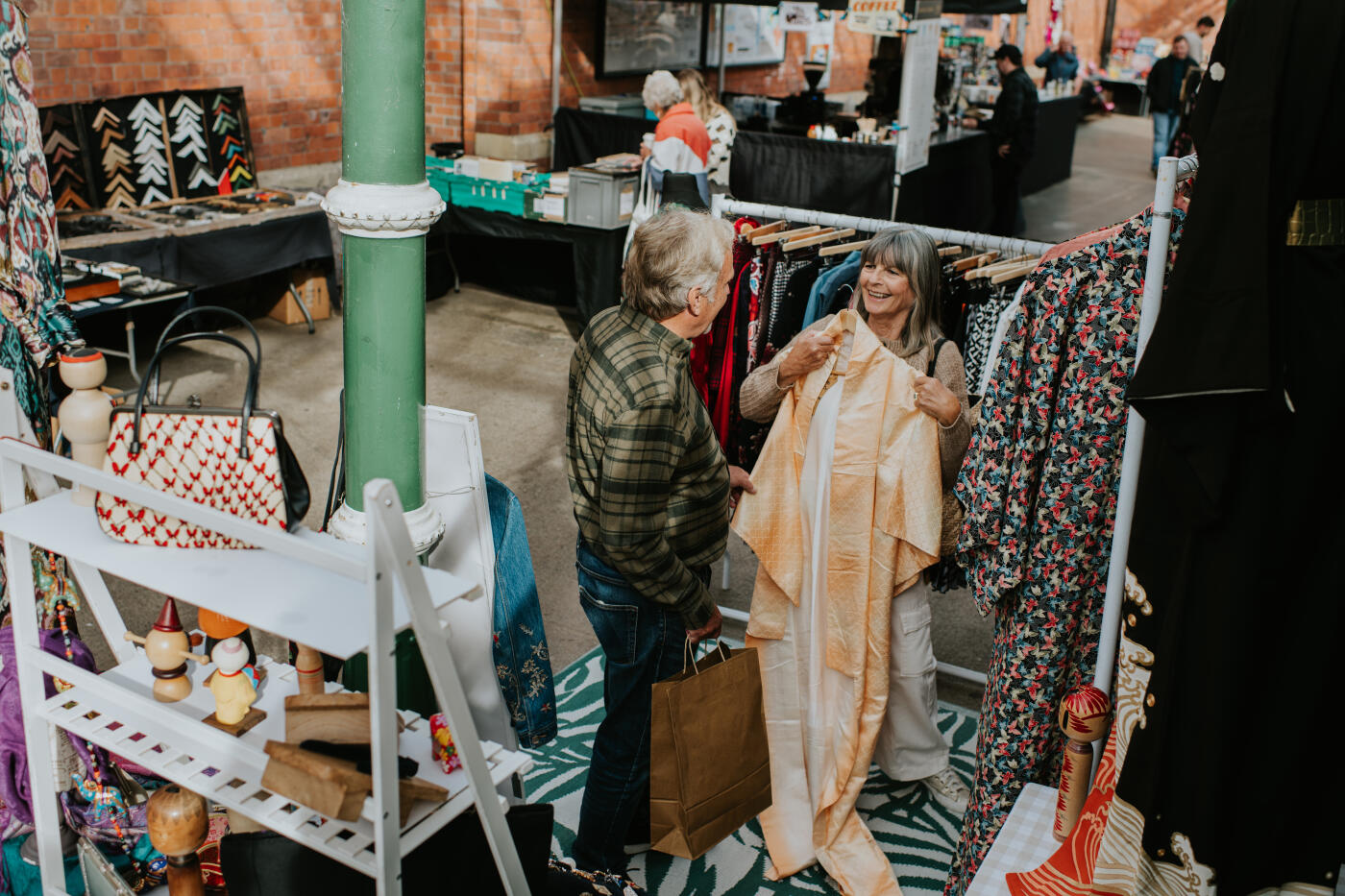 A supportive husband stands close to his wife. The mature couple shop for clothing in an independent boutique in an indoor market. Then woman asks the opinion of her partner.