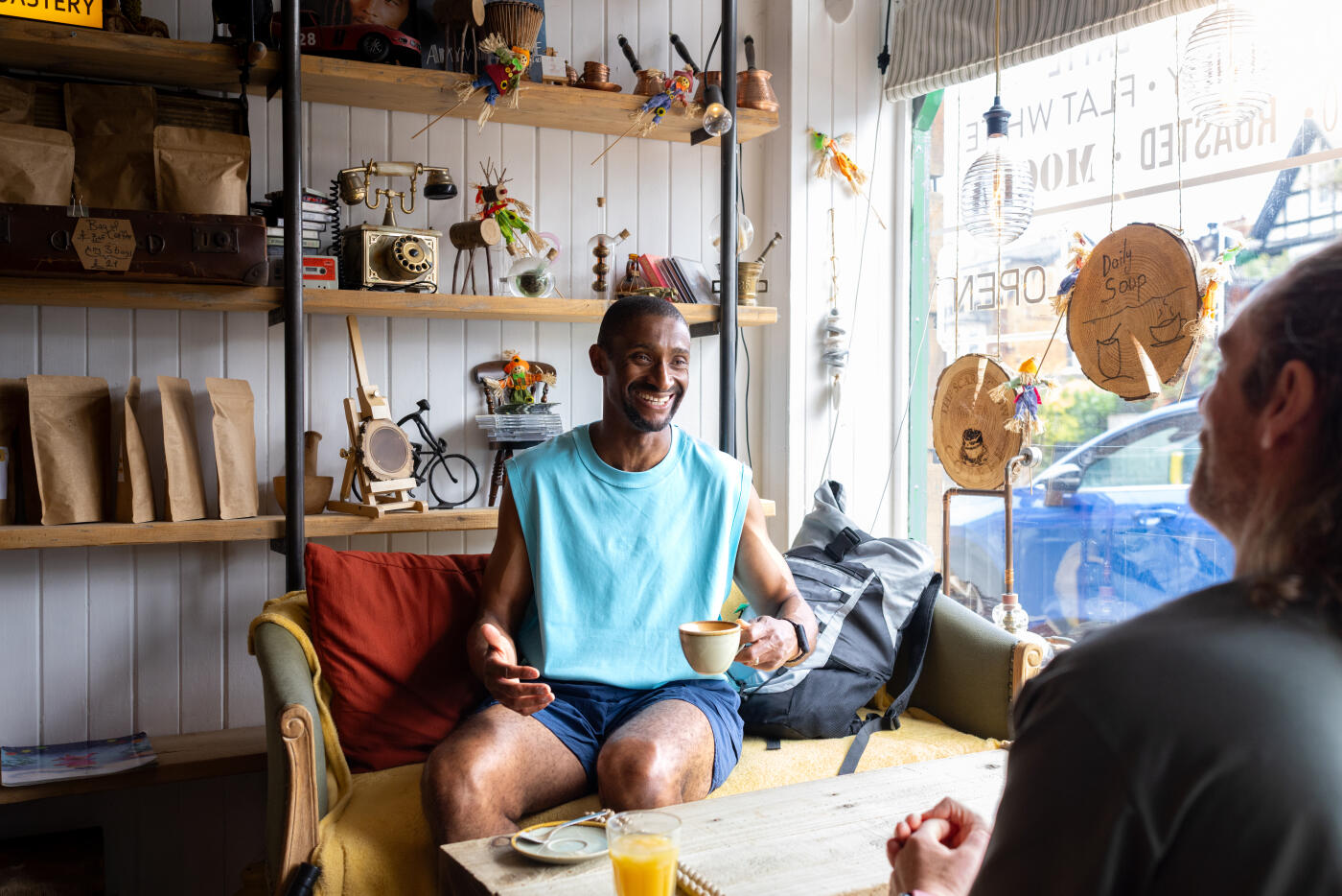 Medium over-the-shoulder shot of two mature adult males relaxing while enjoying a drink in a coffee shop in Monkseaton, North East England. They are sitting face to face on armchairs, talking and smiling, wearing sports clothing.Videos similar to this scenario available.