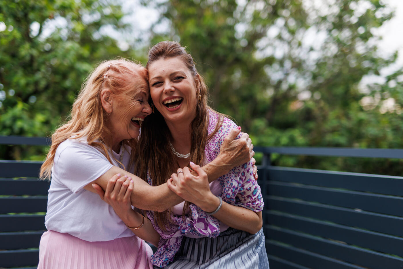 Portrait of mature woman and her mid adult daughter sitting embraced on a balcony, cuddling, having bonding moments. They are laughing together at something funny.