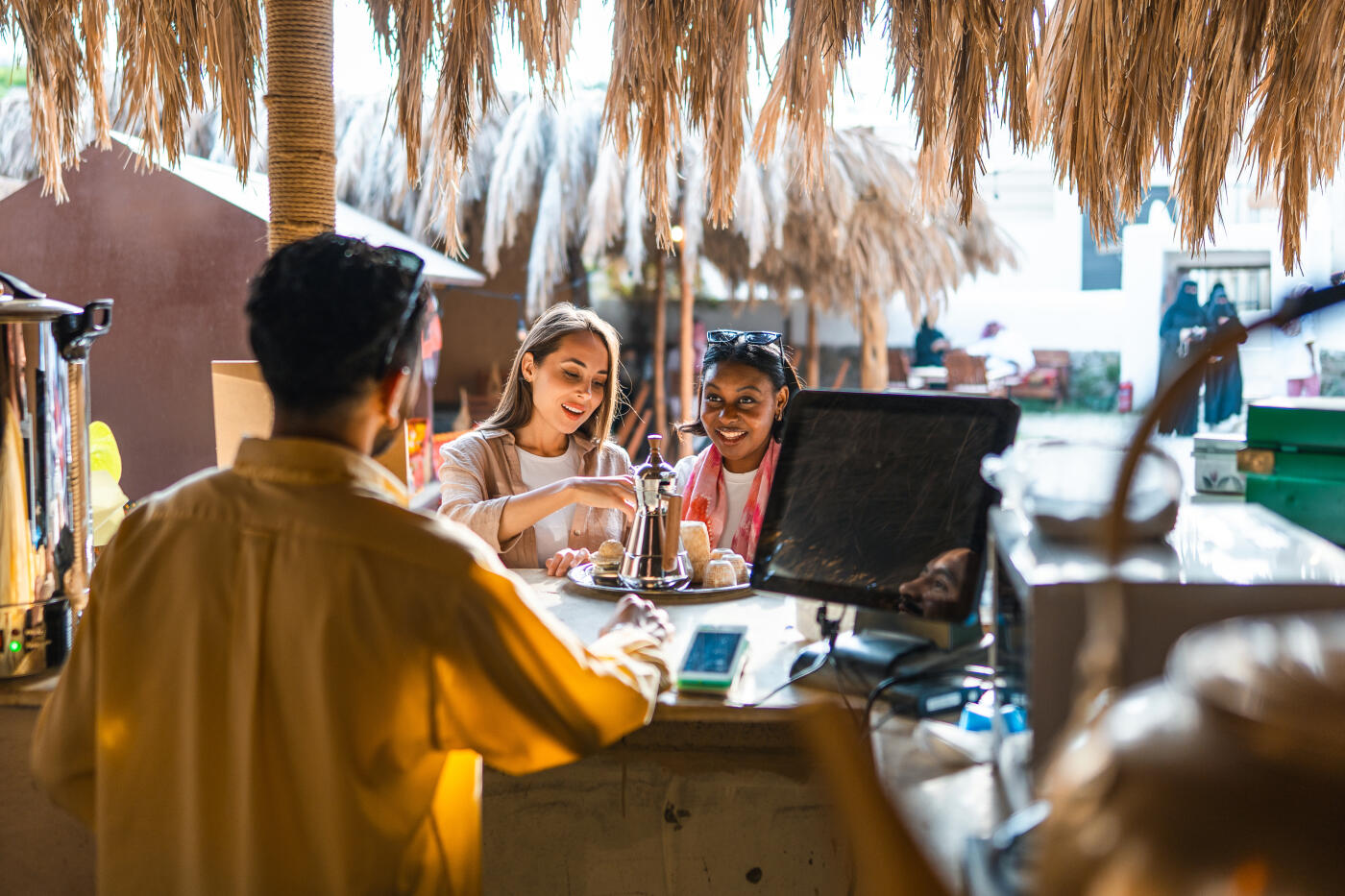Young Caucasian and Arabic females ordering drinks at an outdoor cafe in Muftaha Art Village featuring a relaxed atmosphere with straw canopies and a counter service setting.