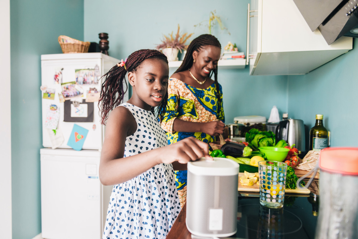 little girl standing in the kitchen and listening to smart speaker together with her mother