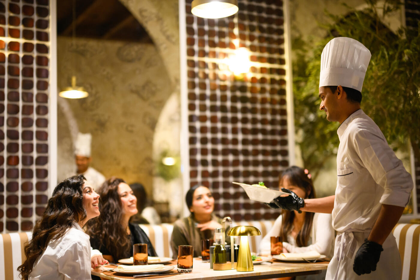 Arab chef personally presenting dishes to a diverse group of women in a luxurious restaurant setting. The elegant dining area is warmly lit, creating a welcoming and sophisticated atmosphere. The chef is dressed in a traditional uniform, engaging with the guests at their table.