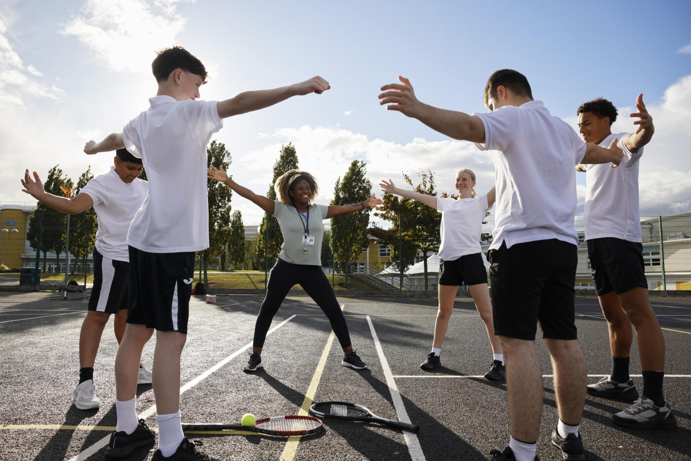 A sports teacher leads a group of secondary school students through a warm-up exercise class outdoors on a sunny day. The students are engaged, smiling, and following instructions, highlighting teamwork, physical activity, and a positive approach to health and fitness in a school setting.
