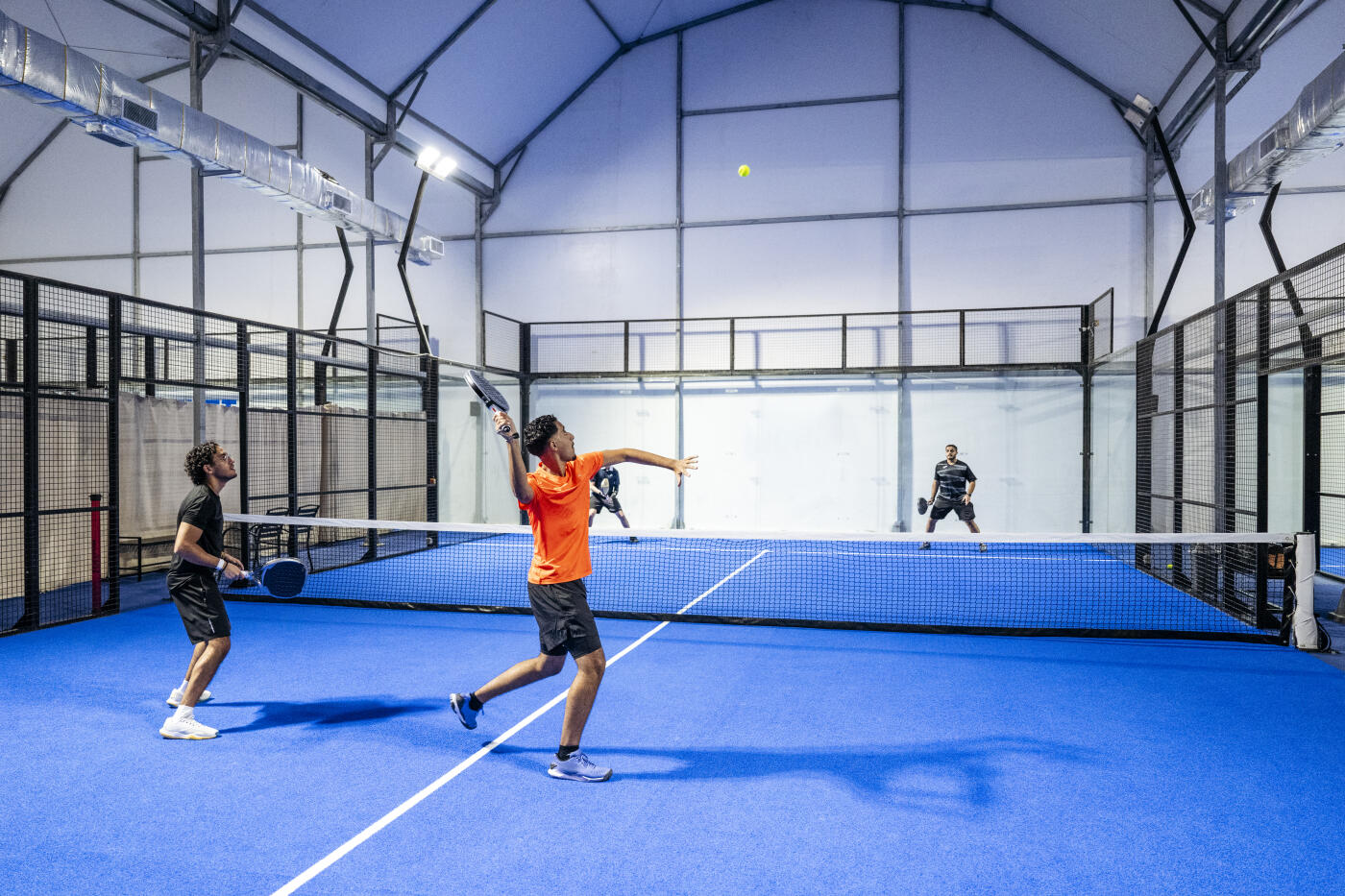 Saudi man about to hit ball during game of padel tennis doubles, on one leg with arm raised