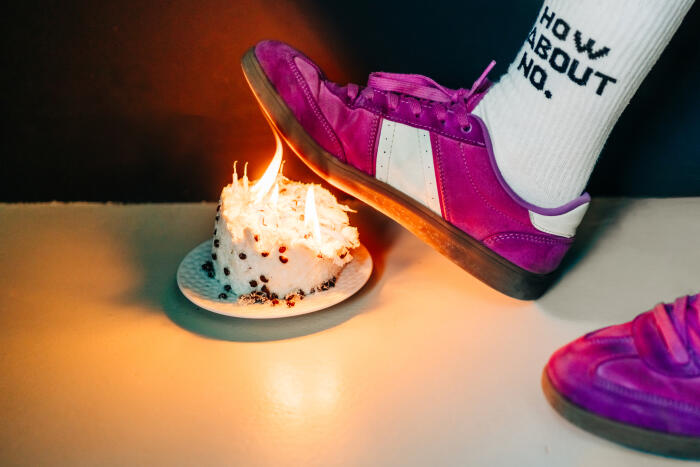 A foot in a vibrant purple sneaker kicks a small, burning birthday cake topped with lit candles.