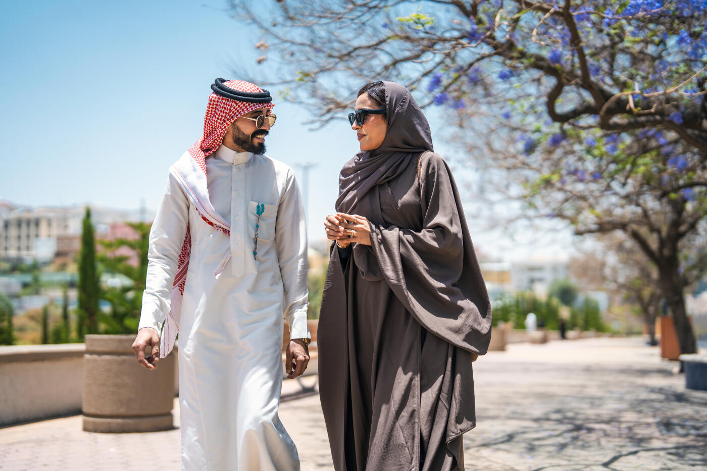 Lovely mid adult Arabic couple walking in Abha public park with Jacaranda trees, enjoying a sunny day and wearing traditional attire featuring vibrant purple blossoms on the trees.