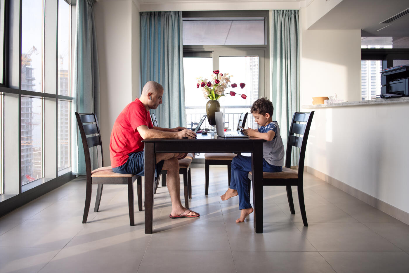 Father and son are busy at dining area. Father is working distantly and child is attending online lessons using laptop and mobile.