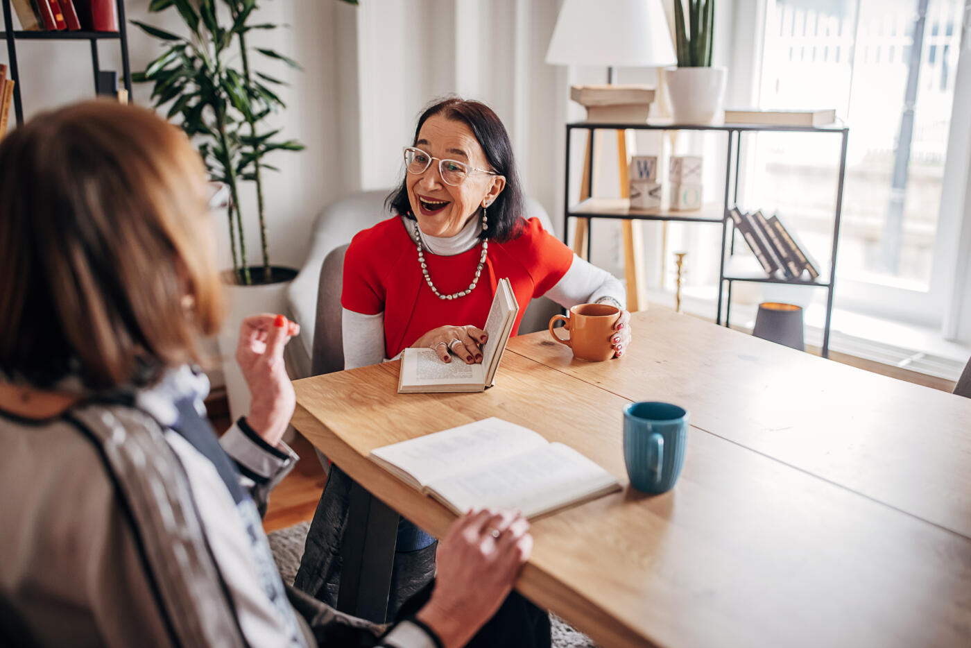 Two senior female friends sitting together at the table at home, reading and discussing books.