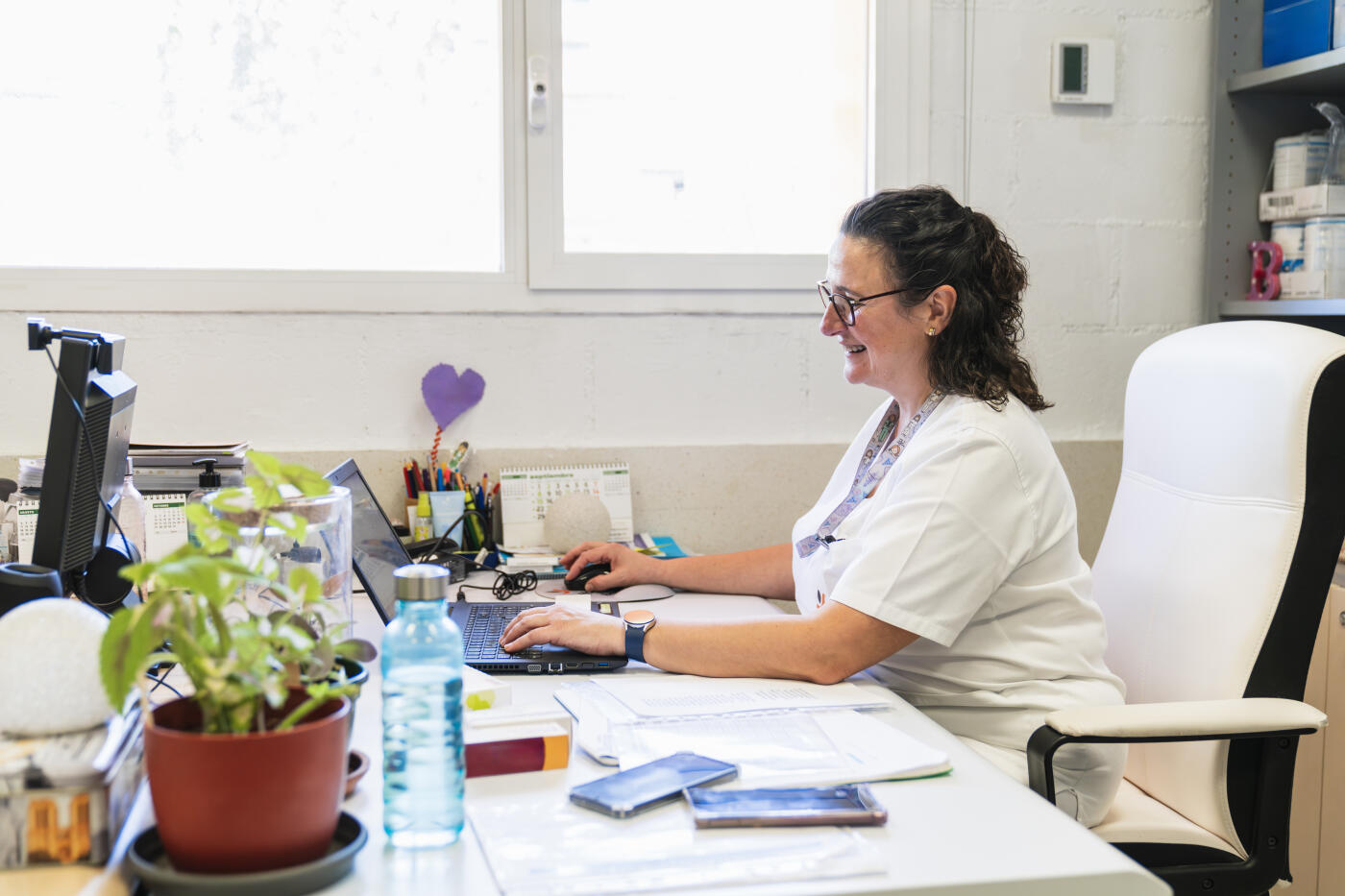 Healthcare worker smiling and typing on a laptop computer, managing tasks in a medical office setting