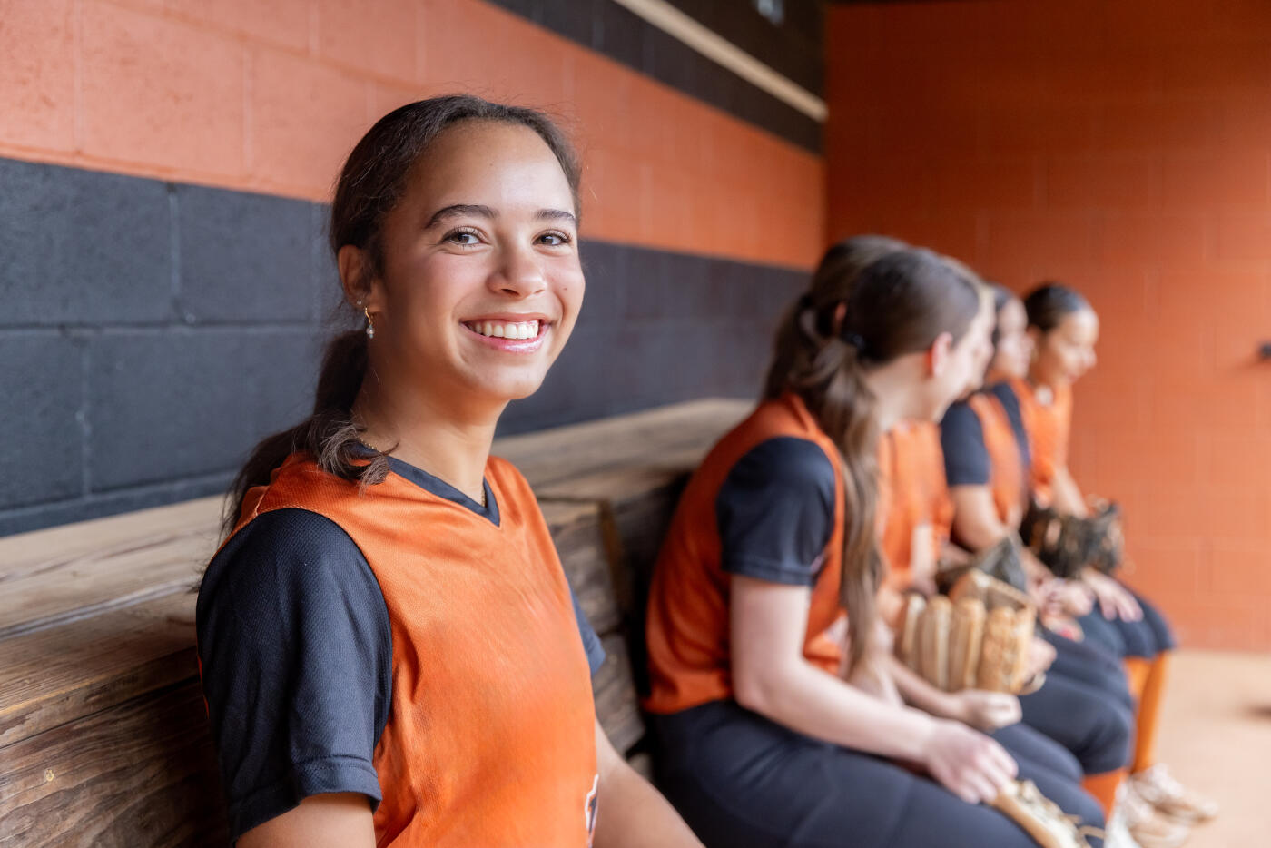 Happy high school age girl is sitting on bench in dugout with her softball teammates before playing game