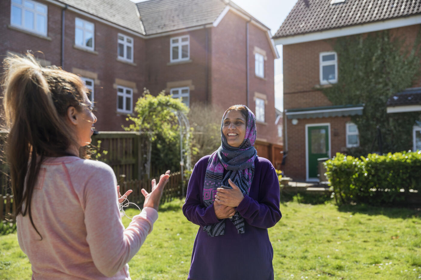 Two female friends standing in a back garden owned by one of them, talking face to face with one another. One woman is wearing a headscarf.