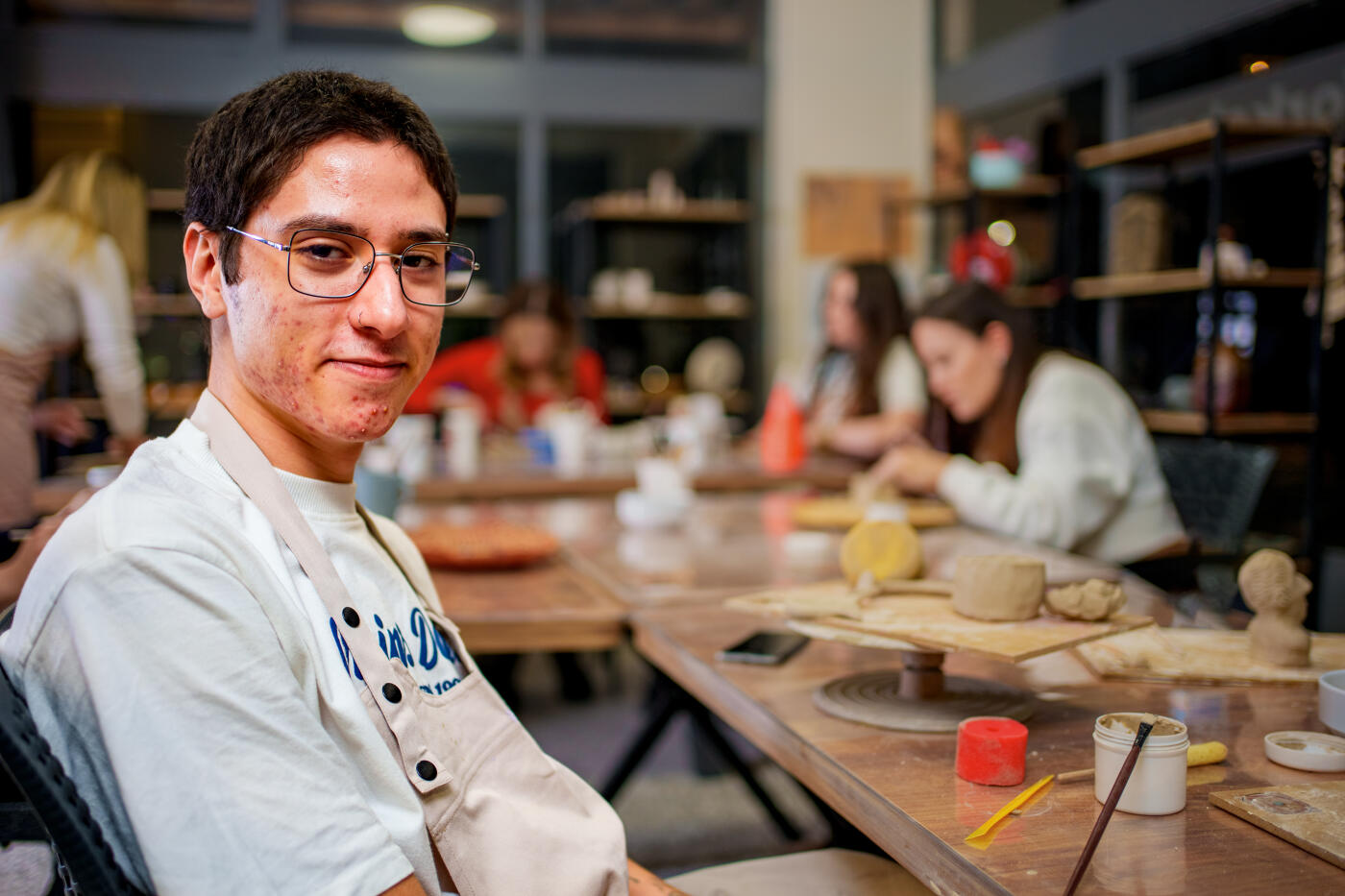 A teenage boy looking at camera and smiling during a pottery class. he is surrounded by other participants, all engaged in creative clay modeling. The setting includes a cozy studio atmosphere with art supplies, sculpting tools, and cups of tea on the table, highlighting the joy of artistic expression and craftsmanship in a community setting.
