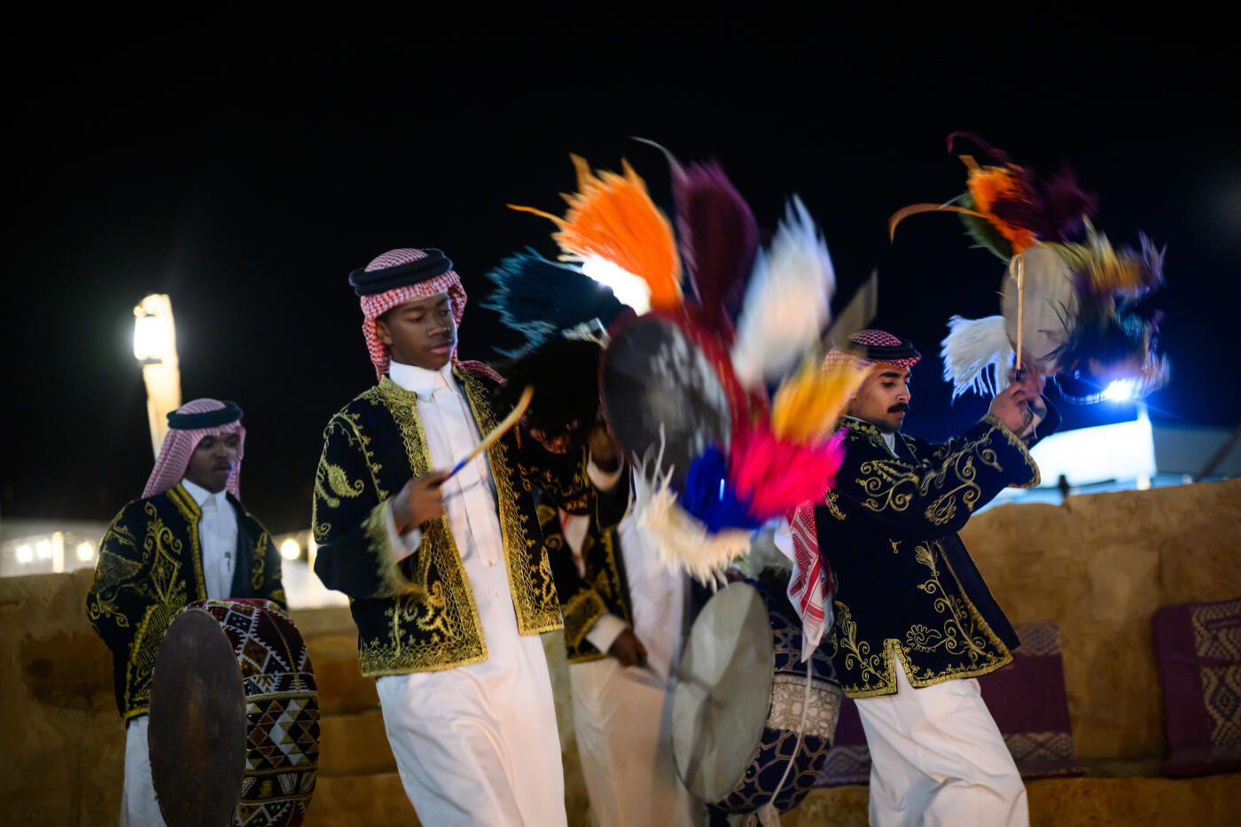 Arab men dressed in ornate traditional attire energetically performing a cultural dance under the night sky. They using colorful feathered props, creating a vibrant and festive atmosphere.
