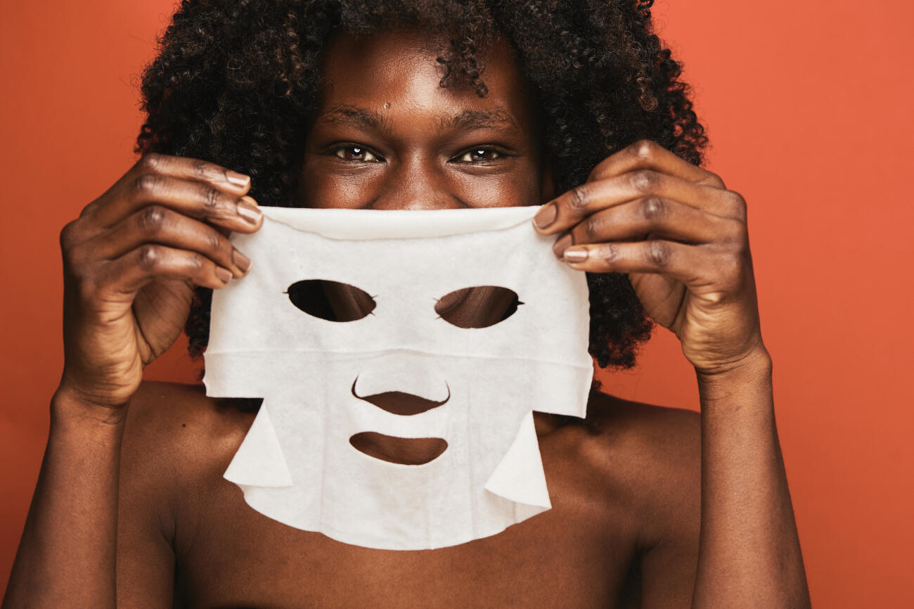 Happy young black woman with curly hair smiles while holding a white sheet mask to her face, enjoying skincare and self-care in a warm terracotta studio close-up