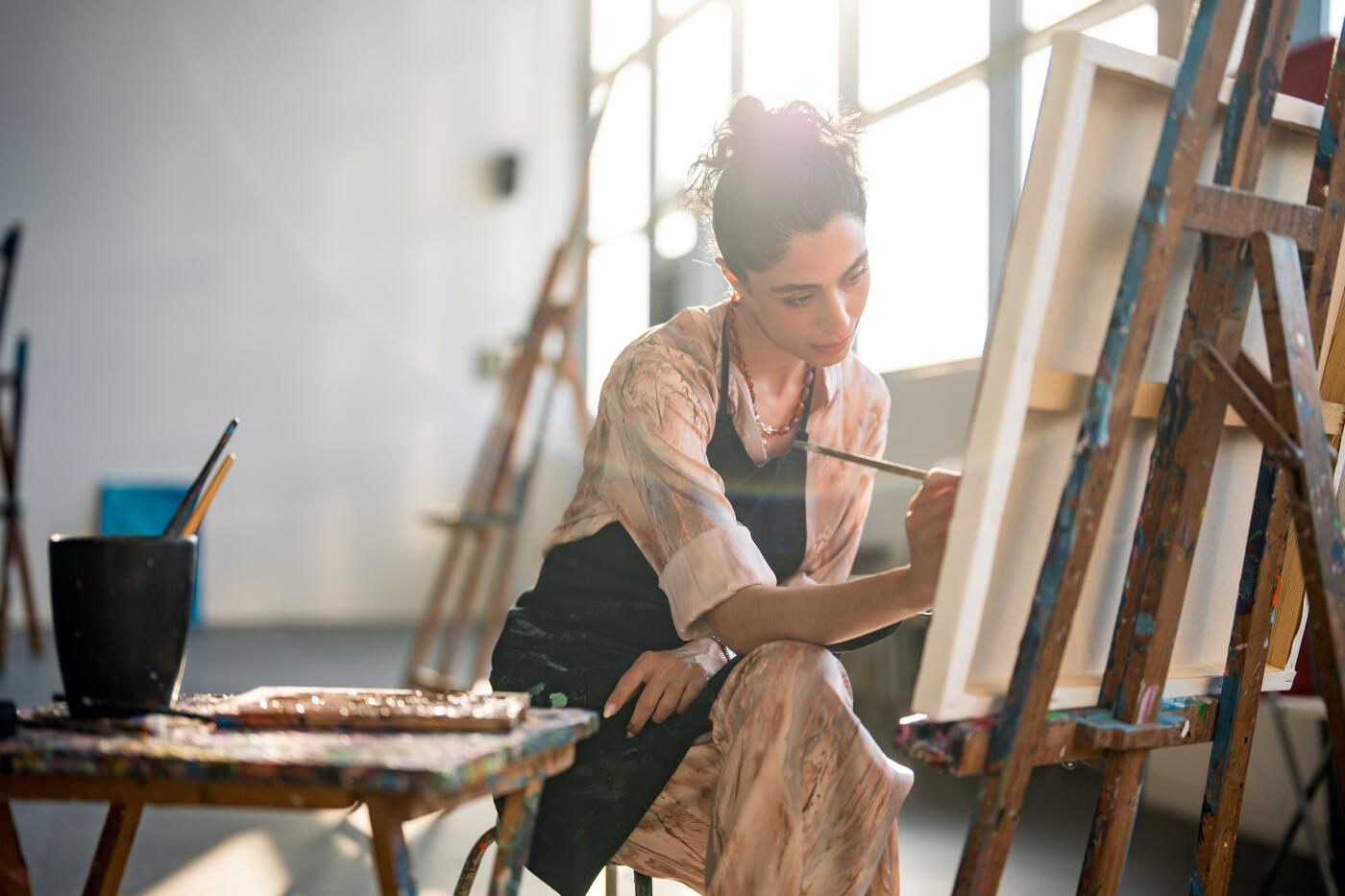 Side-lit portrait of a young Middle Eastern woman artist concentrating on her canvas, working at a wooden easel in a bright, naturally lit studio with artistic tools nearby.
