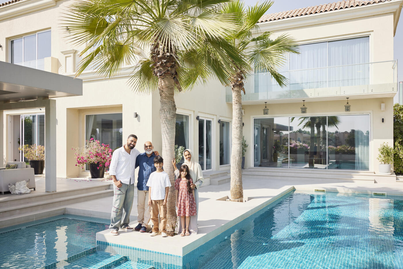 Happy Arabic family by palm tree in front of house. Middle eastern people spending leisure time. They are standing near swimming pool.