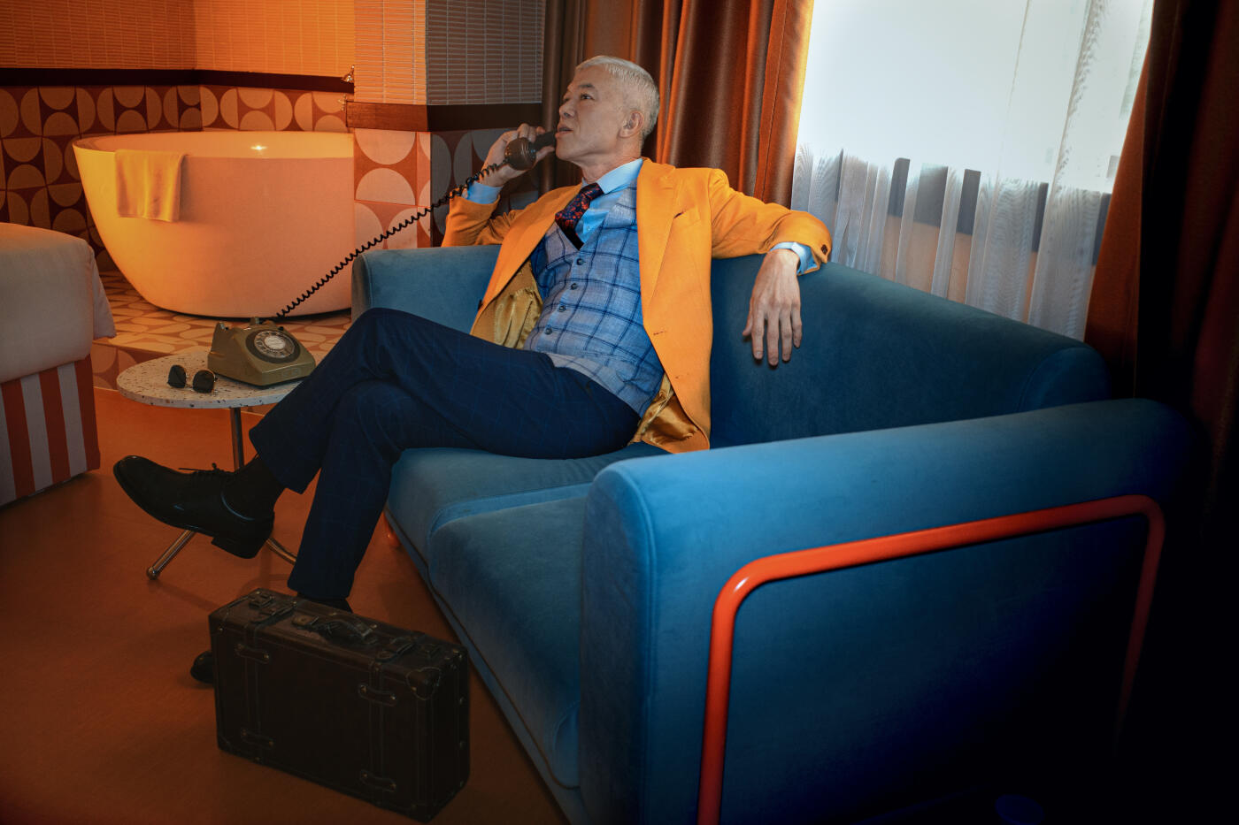 A confident older man in a bright yellow blazer and tailored suit sits on a retro-style blue couch, making a phone call using a vintage rotary phone. With a suitcase at his feet, a freestanding bathtub in the background, and mid-century decor throughout the room, the scene suggests elegance, independence, and a life well-traveled.
