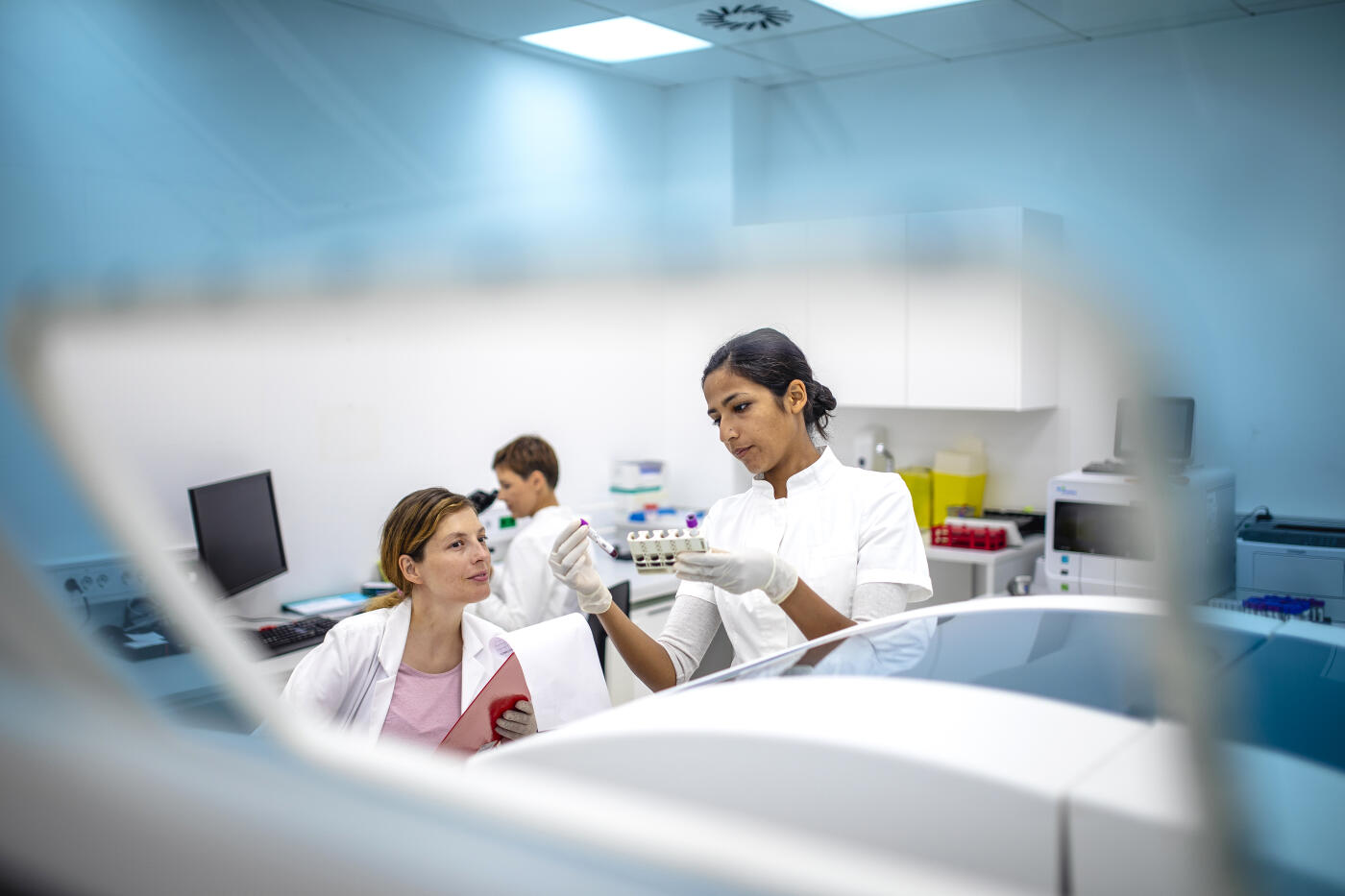 A pregnant Caucasian female scientist collaborates with a young Indian female professional in a bright, modern laboratory, analyzing samples with tools and equipment. Both wear lab coats, emphasizing teamwork and scientific expertise.