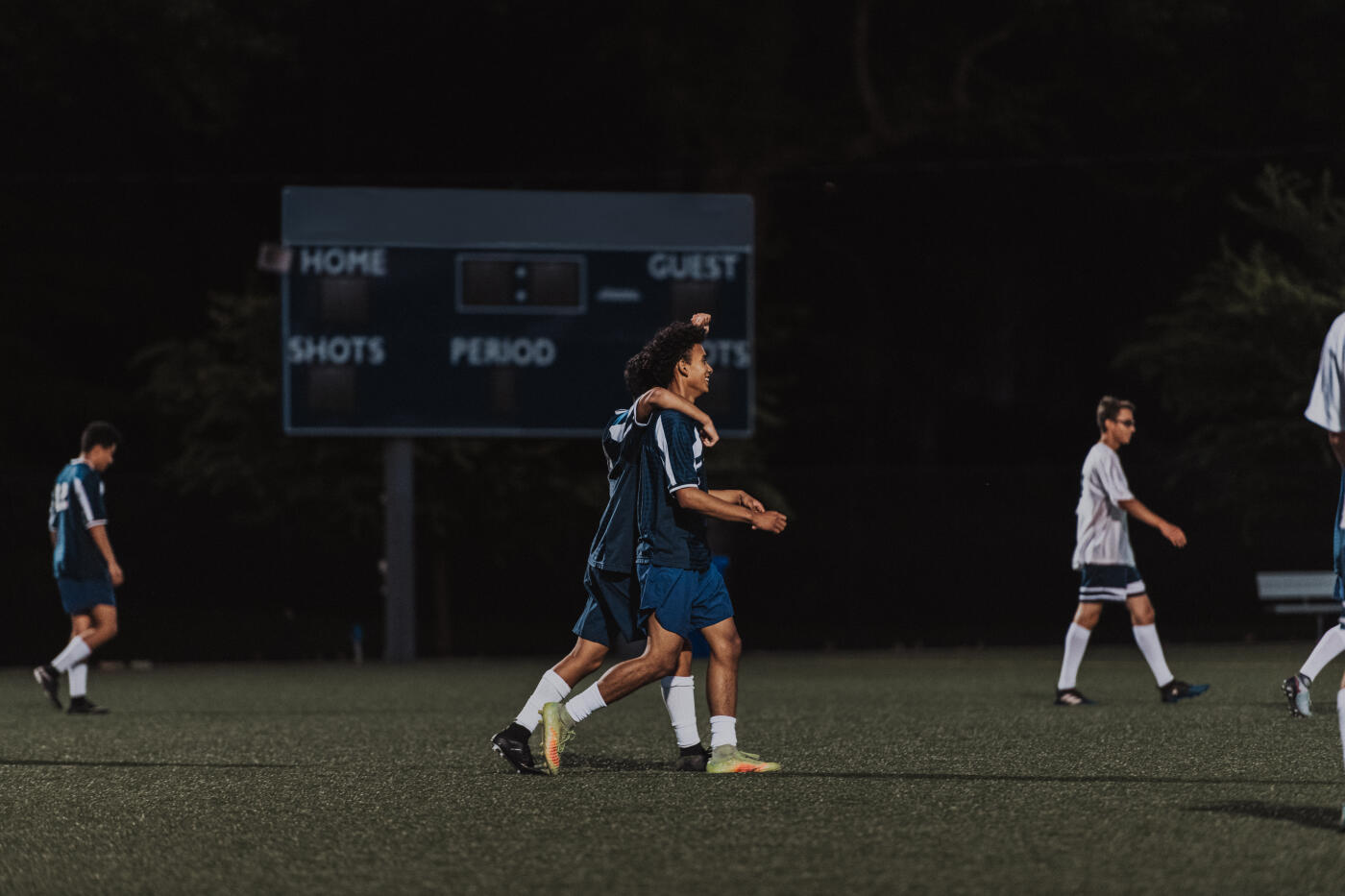 A group of young adult men walk on a turf field at night while playing against each other in a competitive soccer game.
