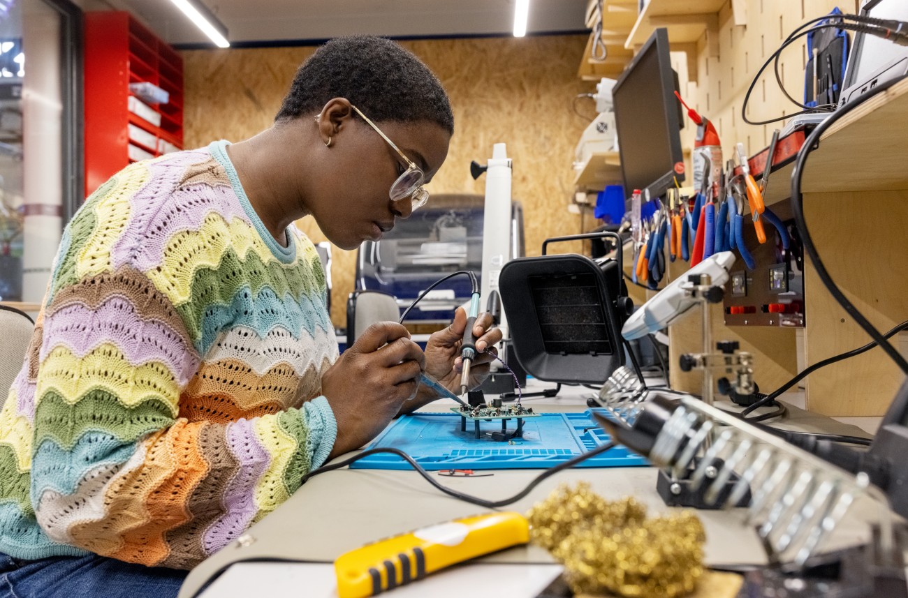 Side view of African woman repairing circuit board at makers space. Female electrical engineer soldering electronics circuit board with a soldering iron at workshop.