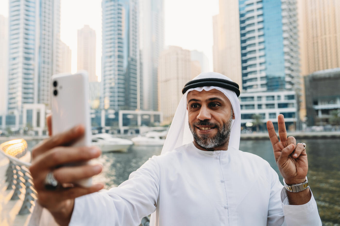Emirati businessman smiling while taking a selfie and making the peace sign, surrounded by the stunning Dubai Marina cityscape