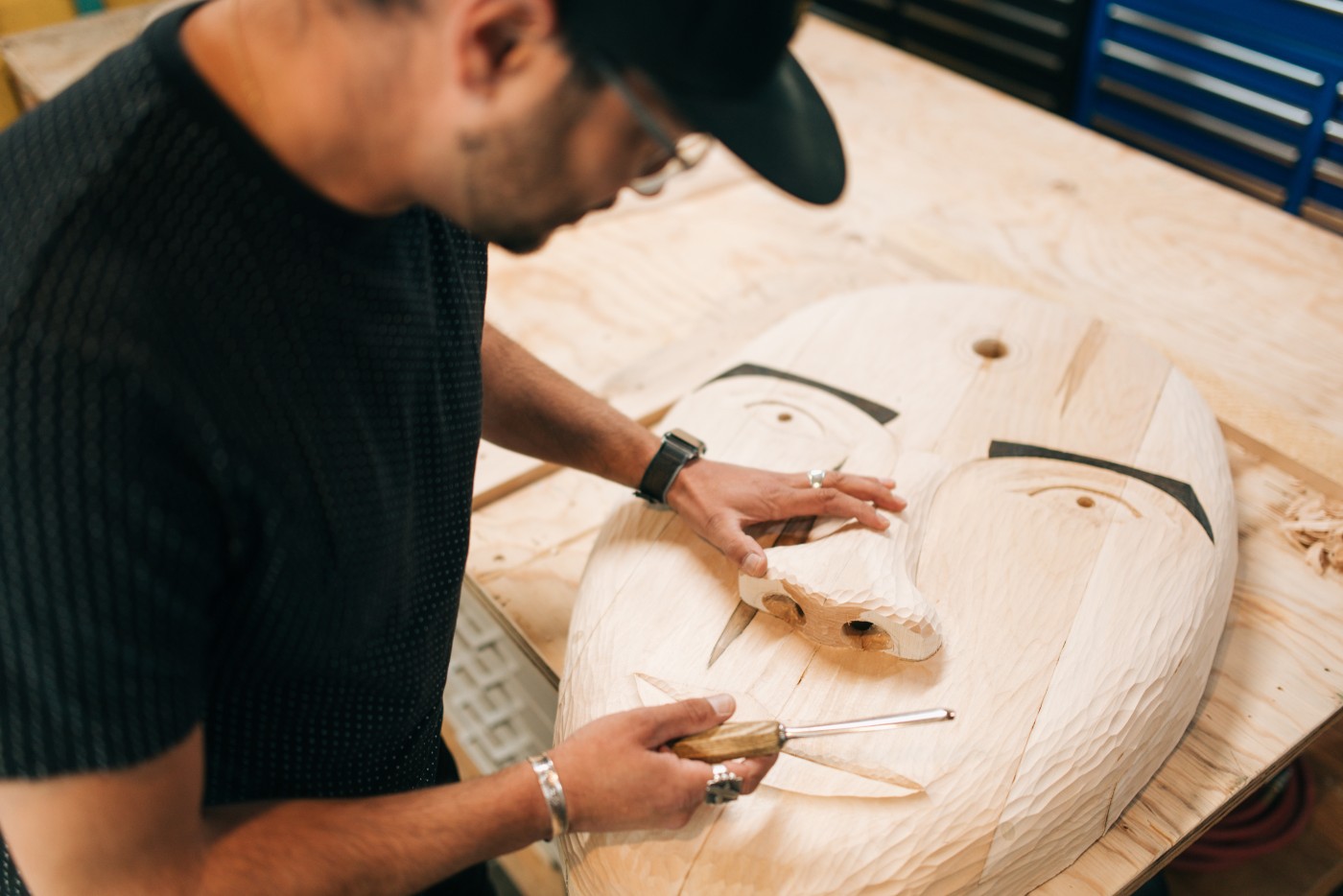 A multiracial Alaskan man in his workshop, planning, carving, and networking online with his hand carved wooden masks.  Traditional culture with a modern twist.