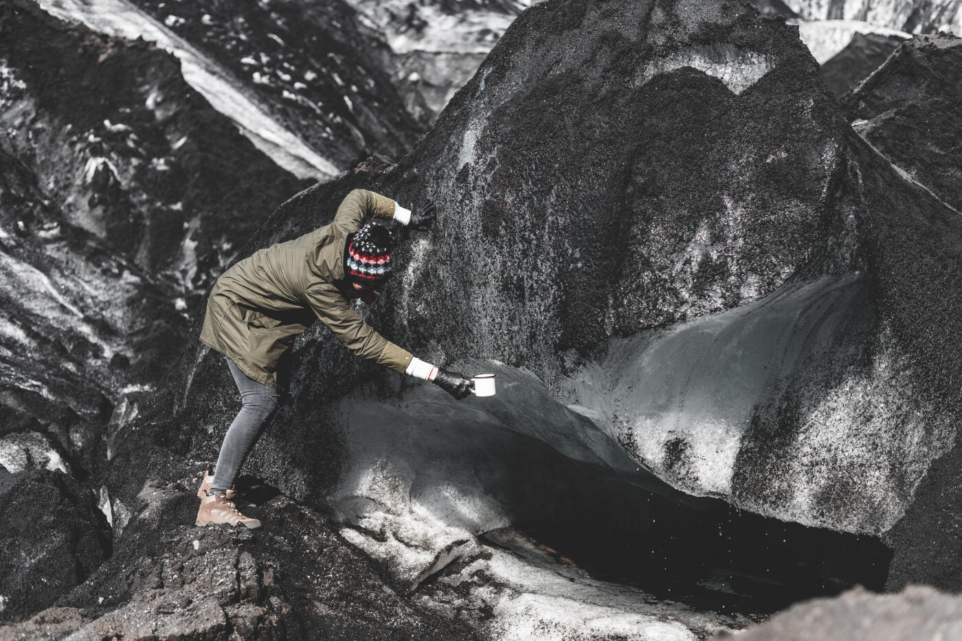 A girl in a khaki jacket surrounded by ice and black sand from the glacier collects water from it into a metal white mug.
