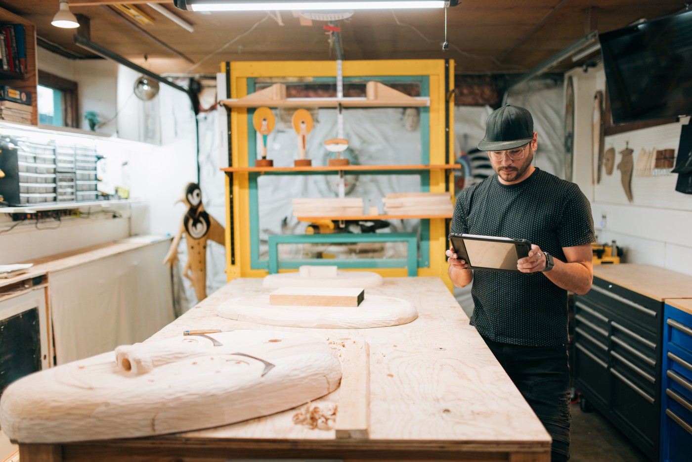A multiracial Alaskan man in his workshop, planning, carving, and networking online with his hand carved wooden masks.  He lists pieces of art online with a digital tablet computer. Traditional culture with a modern twist.