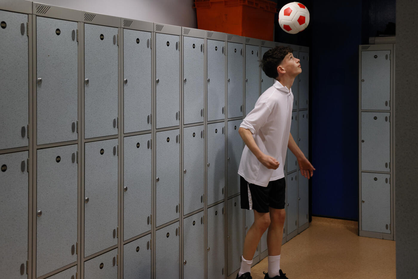 A young male secondary school student practises his football skills alone in an empty locker room. The image highlights focus, determination, and personal dedication, capturing concentration and skill development in a quiet school environment.