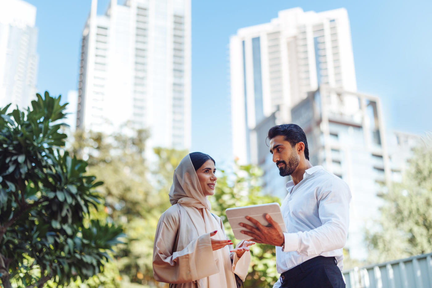 A young Caucasian female in a beige hijab and a Middle Eastern male in a white shirt discuss plans using a digital tablet outdoors in Dubai.