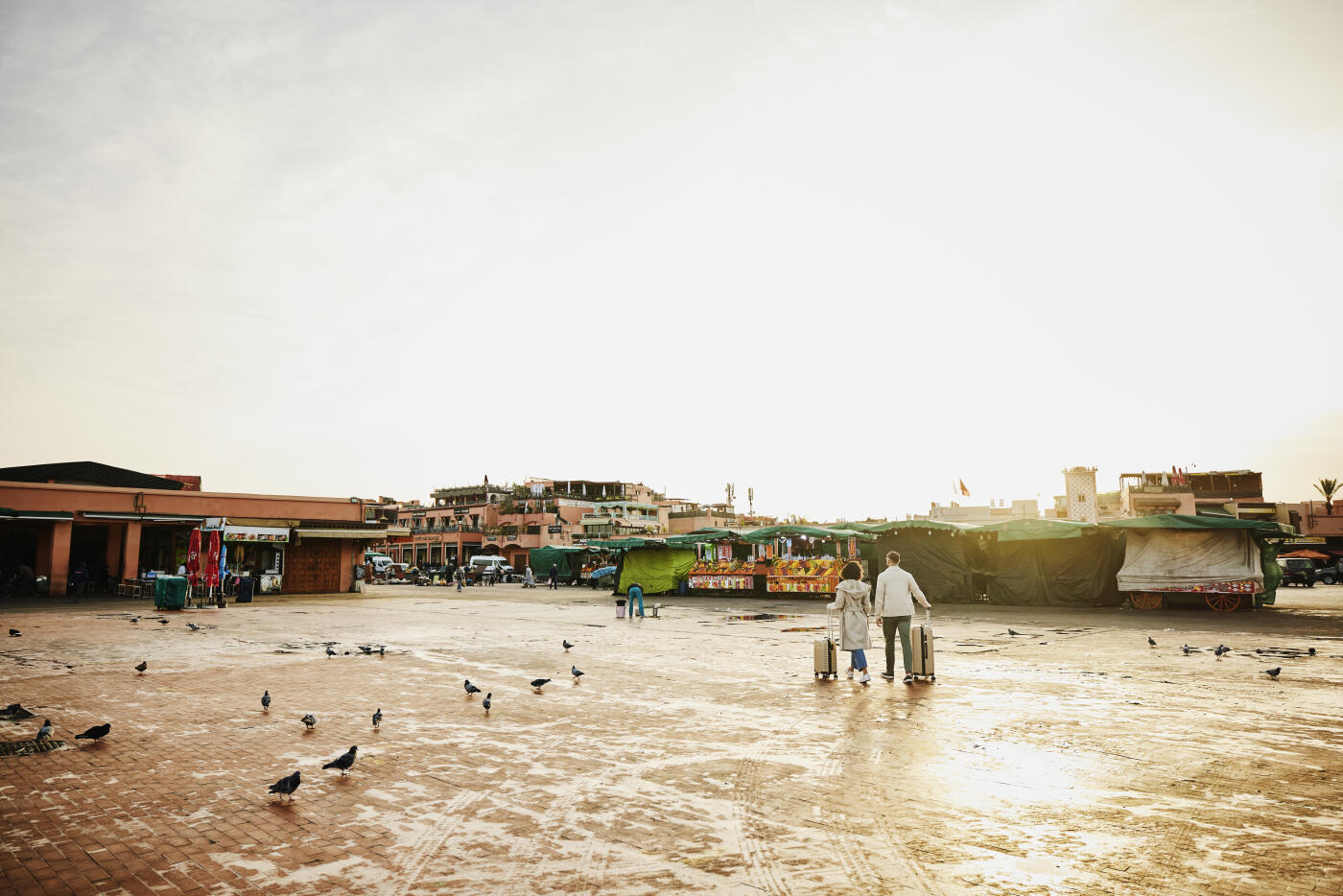 Extreme wide shot rear view of couple with rolling luggage walking across Jemaa el-Fna square at sunrise during vacation in Marrakech