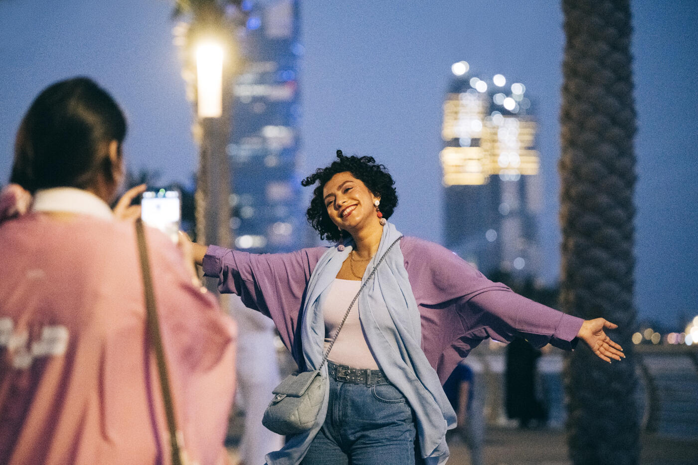 Candid portrait of woman holding smartphone and taking photo of attractive young woman in her 20s with cheerful carefree expression and arms out wide, city lights in background