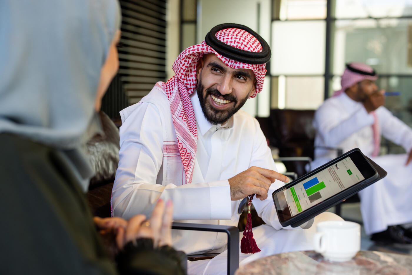 Saudi businessman holding a digital tablet while talking with a female colleague sitting at a café. Arab business professional pointing at statistical data on the tablet and discussing work with a businesswoman.
