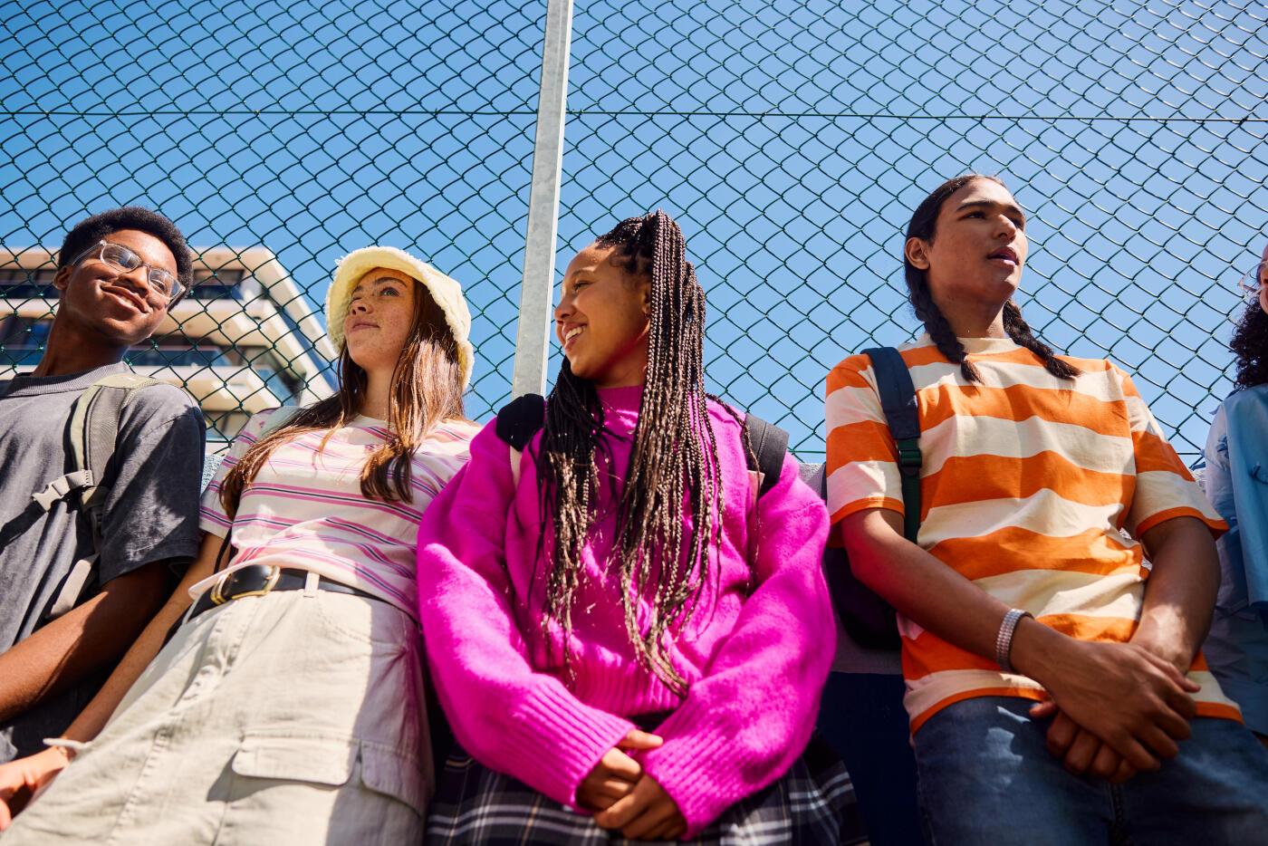 Four diverse high school students are hanging out by a fence, chatting and enjoying each other's company after school