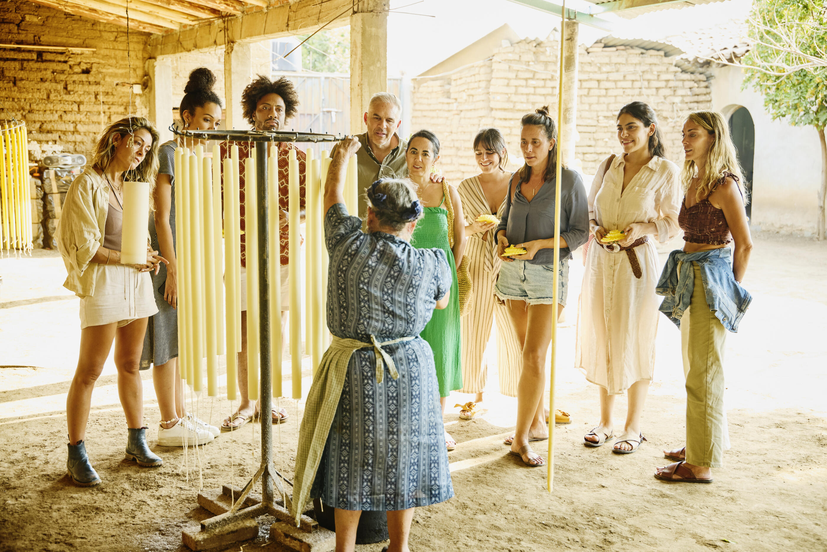 Wide shot of curious tourists learning from a master female candlemaker while she crafts tall ceremonial candles during an outdoor studio tour