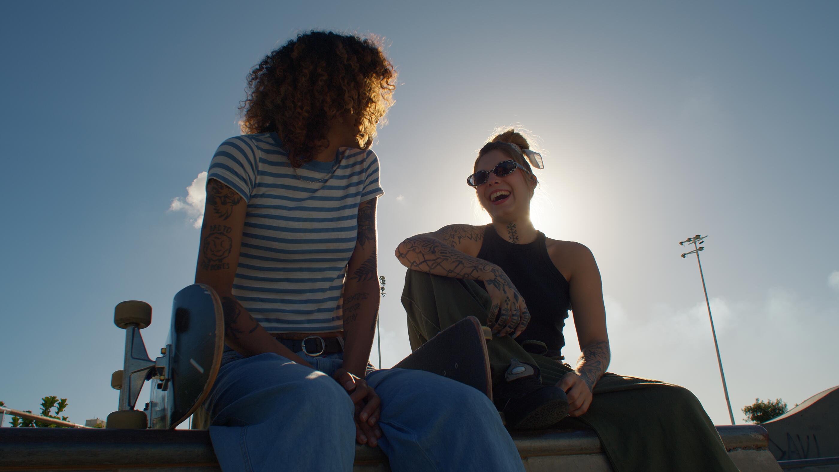 Portrait of two female skateboarders sitting together at a sunny outdoor skatepark, smiling and holding their skateboards.