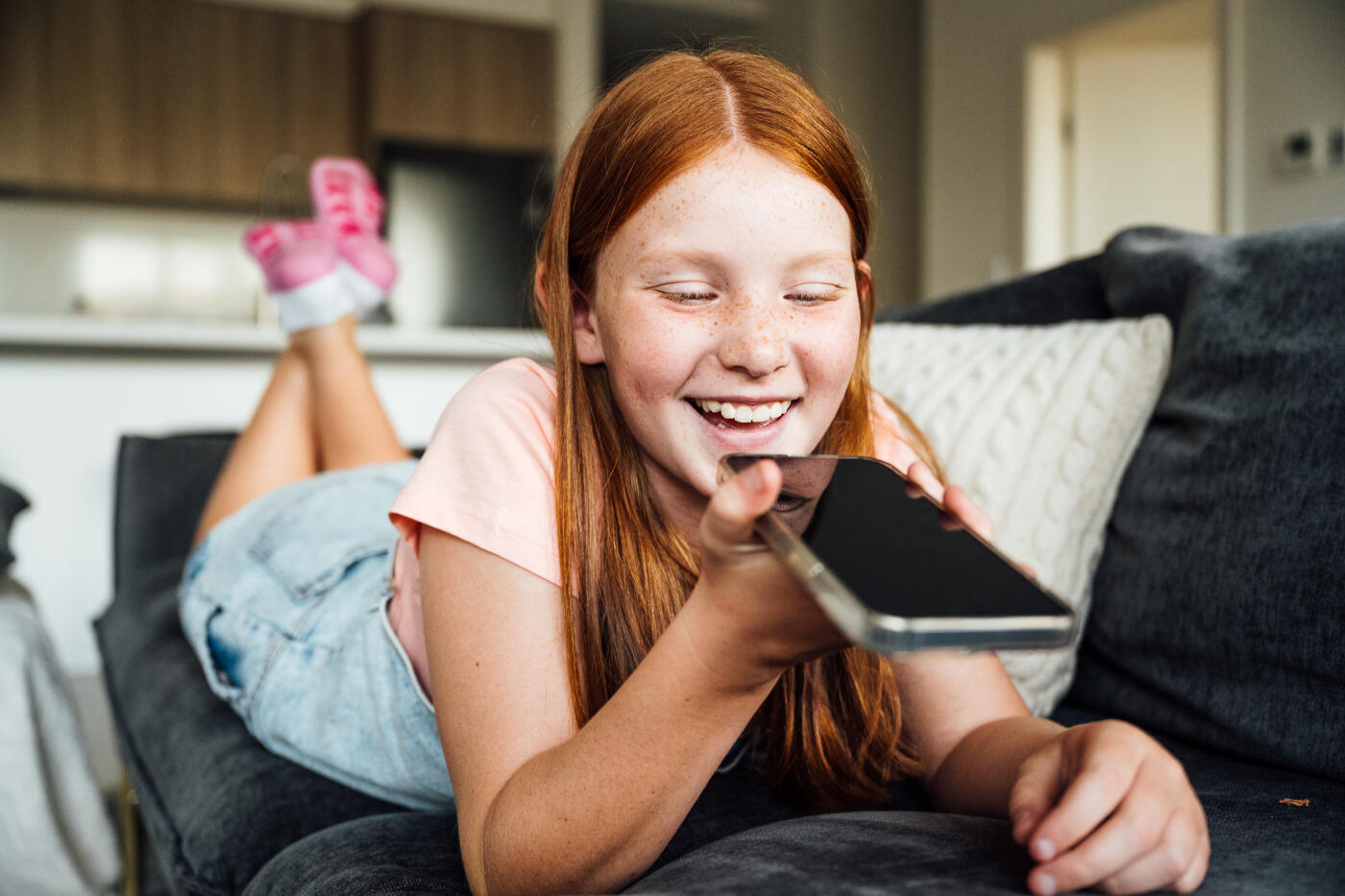 Smiling teenage girl with red hair and freckles sitting on a couch in a modern home setting wearing casual clothes and using smart phone