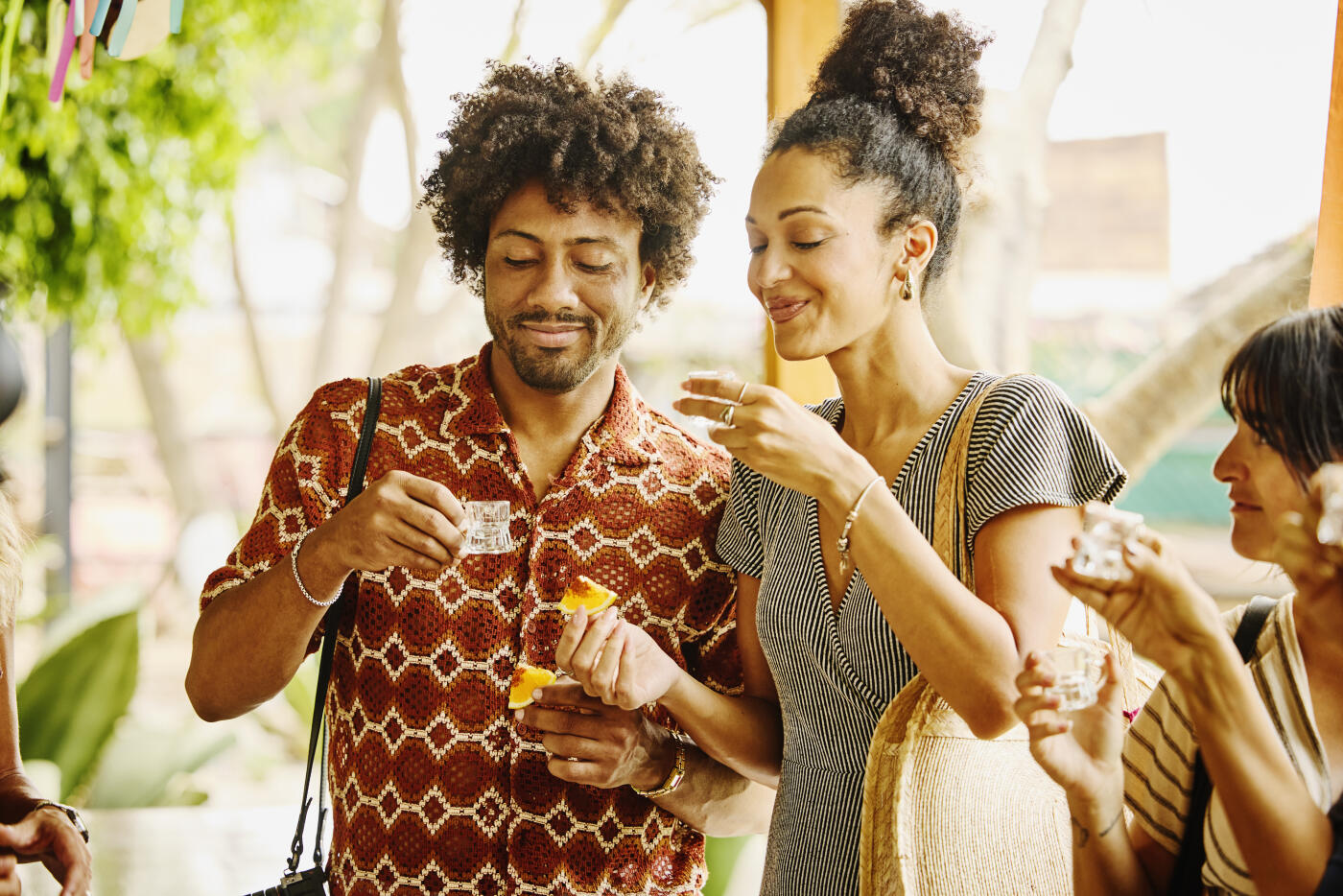 Medium shot of smiling couple raising shot glasses and preparing to taste artisanal mezcal with traditional orange slice garnish during tasting tour