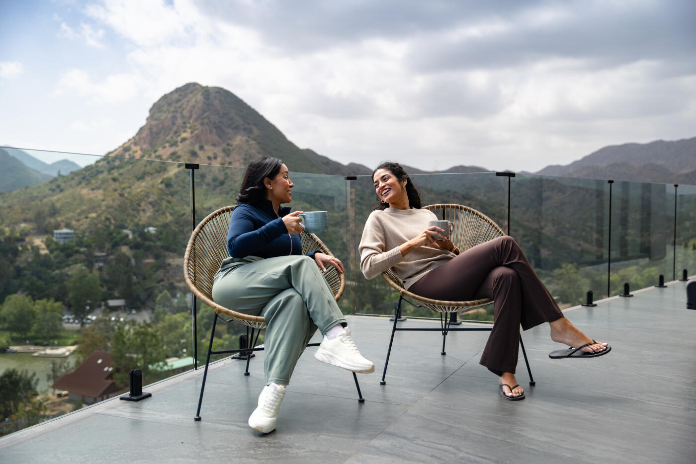 Two mid adult women sit in woven lounge chairs on an outdoor terrace with a glass railing, holding ceramic mugs and talking during the day. Rolling hills covered with green vegetation rise in the background beneath an overcast sky in the Agoura Hills area of Southern California. The relaxed setting captures a warm moment of friendship and conversation.
