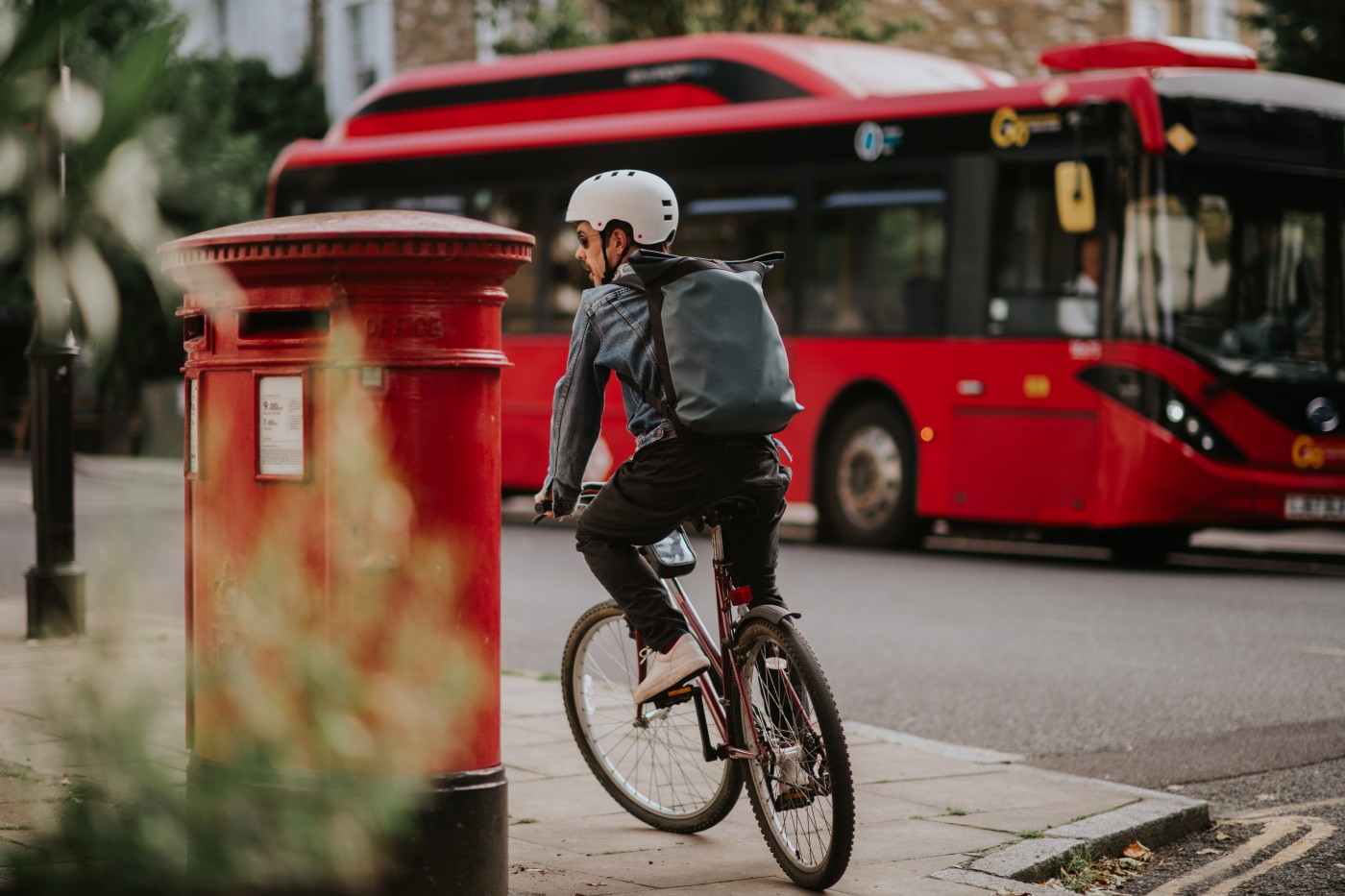 A man cycles on a footpath in a UK city. The scene is very London-centric with a recognisable red post box and big red bus in the background. Depicts commuting through an urban environment.