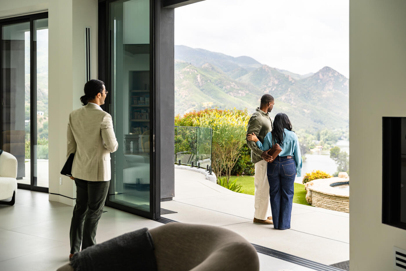 A mid adult African American couple stands on a concrete patio with their arms around each other, looking out over rolling green mountains and a lake in the distance. A Middle Eastern real estate agent in a light blazer holds a folder and stands inside the open-plan living room near a sliding glass door in the morning. The luxury home features floor-to-ceiling glass walls, a glass balcony railing, landscaped gardens, and a stone-edged spa on the terrace overlooking the valley in Agoura Hills, California.